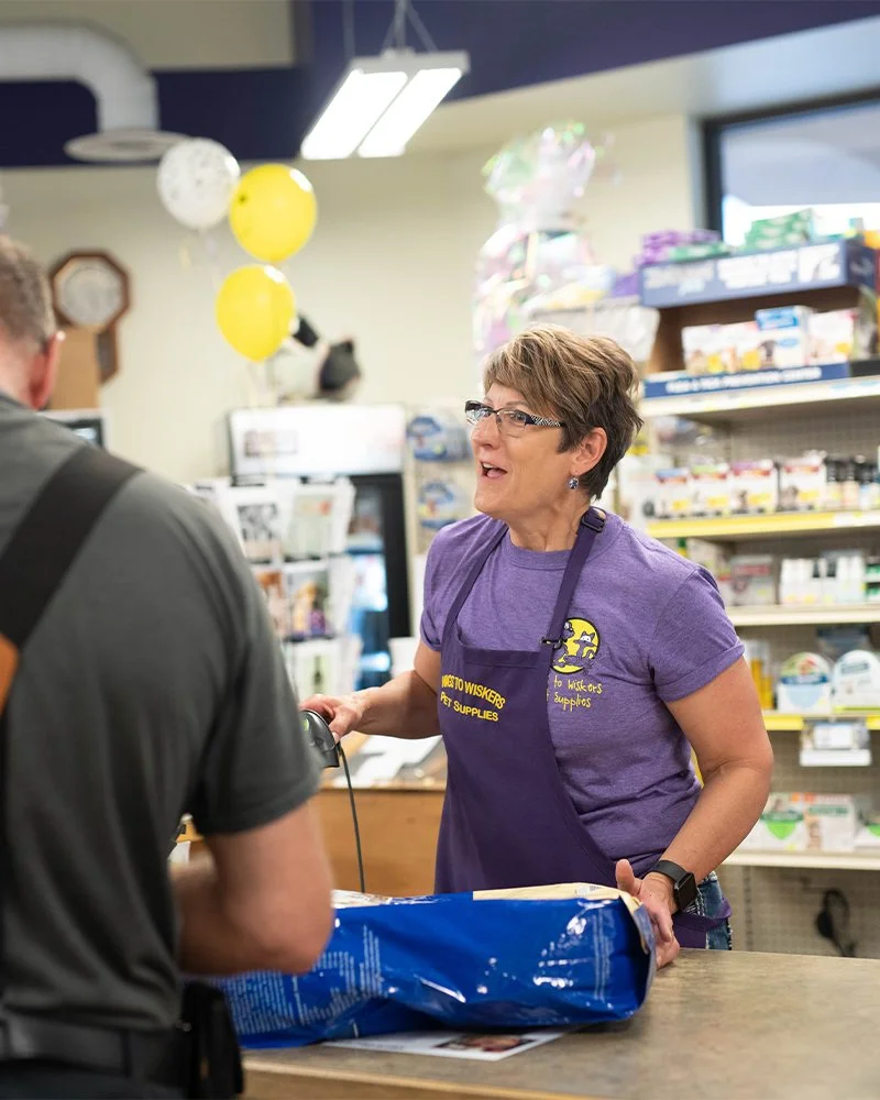 A woman wearing glasses and a purple apron talking to a customer at a checkout counter inside a store with balloons and shelves of products in the background.