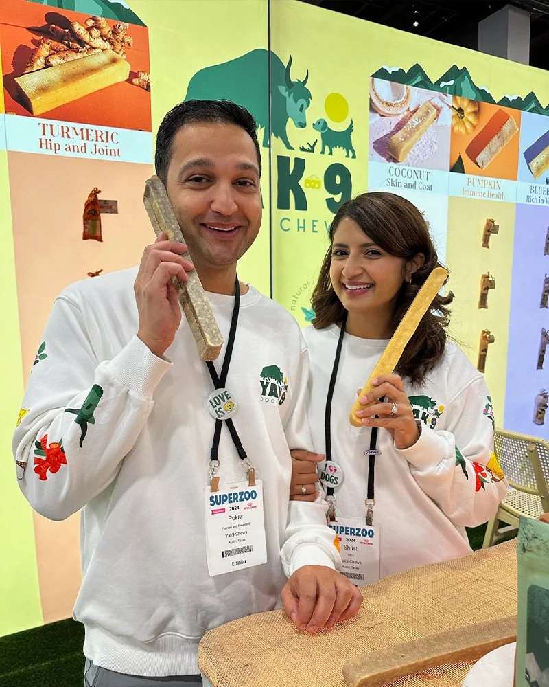 Two smiling people holding turmeric sticks at a health product booth, with a colorful backdrop showing various natural ingredients and benefits.