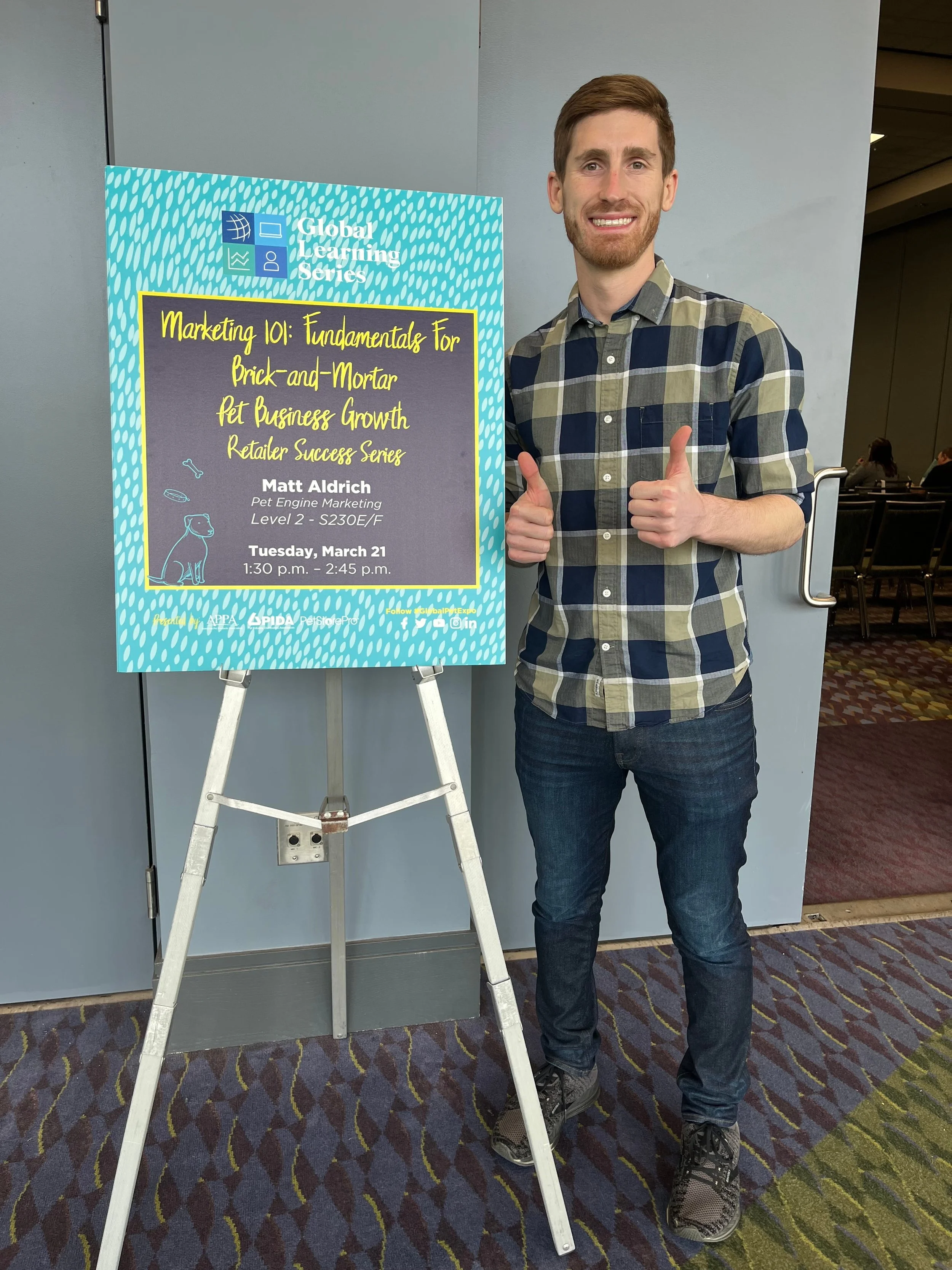 A man standing next to a sign at a conference, giving a thumbs-up and smiling, in a patterned plaid shirt, dark jeans, and sneakers.