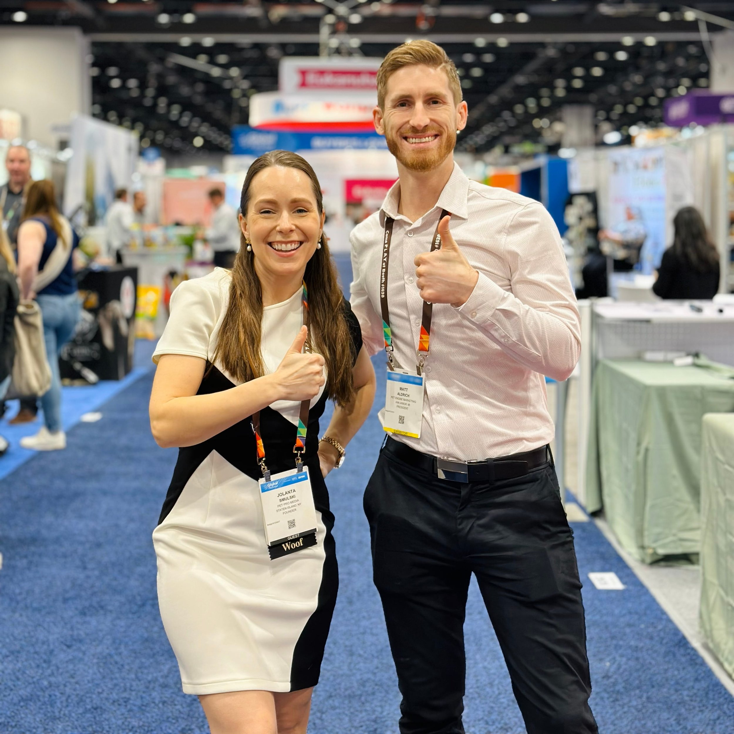 Two smiling people, a woman and a man, standing at a trade show or conference, giving thumbs-up gesture. They are wearing conference badges and lanyards, with booths and other attendees in the background.