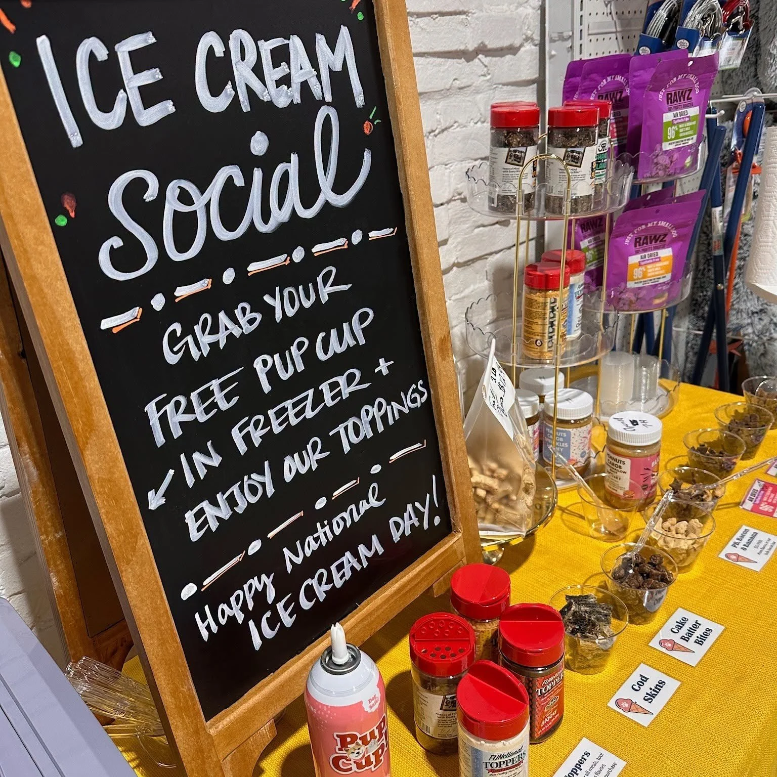 A blackboard sign with hand-written white text advertising free ice cream in a cup or cone, topped with various toppings. Nearby, a display of jars and containers with different toppings, some labeled like 'Cake Batter Bites' and 'Candy Sprinkles,' is set up on a yellow tablecloth.