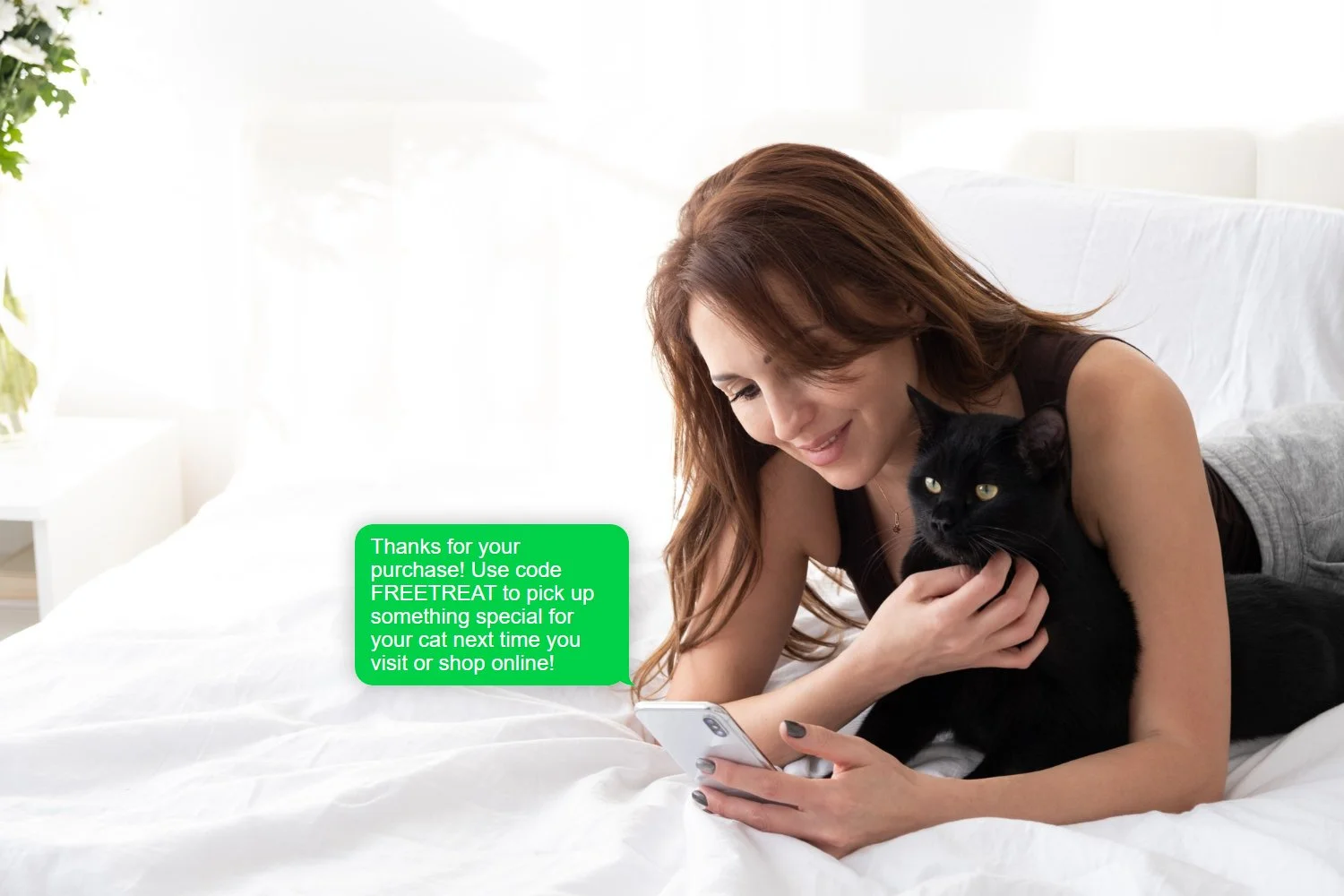A woman with long brown hair lying on a bed, smiling while holding a black cat in her arms and looking at her smartphone. There is a green text message bubble on the image that reads: 'Thanks for your purchase! Use code FREETREAT to pick up something special for your cat next time you visit or shop online!'