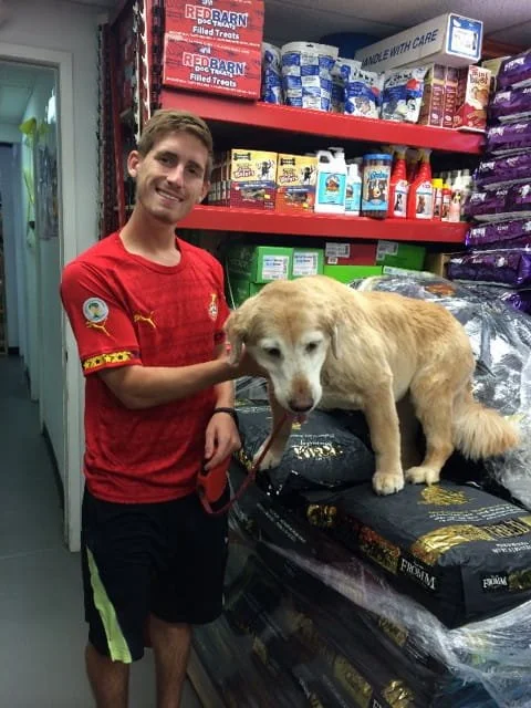 A young man in a red soccer jersey is standing in pet store aisle, smiling, while holding a leash attached to a large cream-colored dog sitting on a bag of pet food on display.