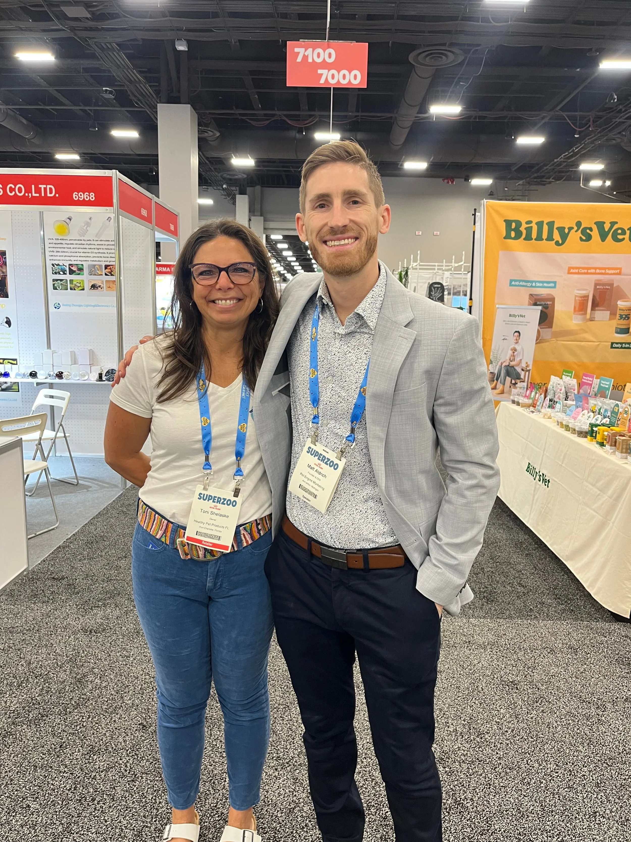 Two people, a woman and a man, standing side by side at a trade show or convention, smiling at the camera. They are wearing name badges and lanyards, with booths and displays visible in the background.