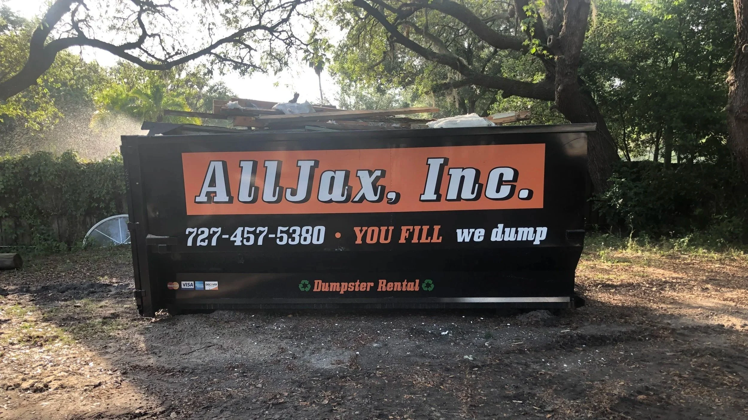A black dumpster with an orange sign reading "AllJax, Inc." and a phone number, providing dumpster rental services with the slogan "You Fill, We Dump" and logos of payment methods, surrounded by trees and outdoor scenery.