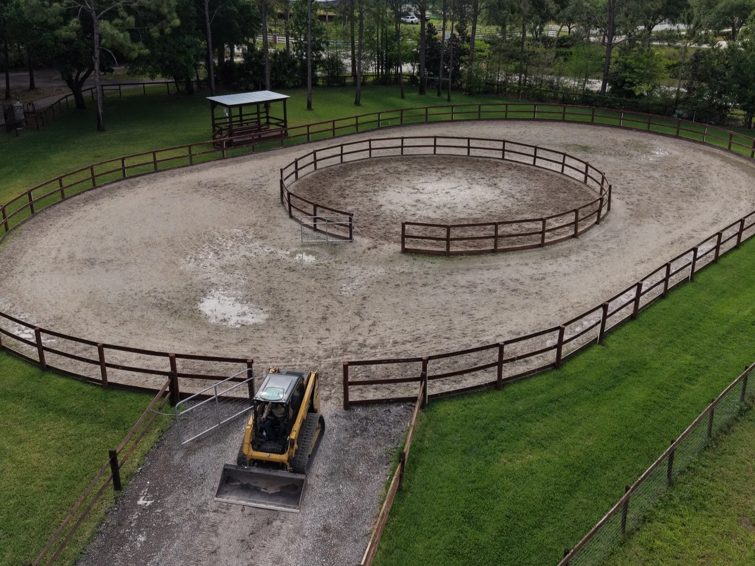 A dirt riding arena enclosed by wooden fences, with a small vehicle in the foreground, and trees and a small shelter in the background.