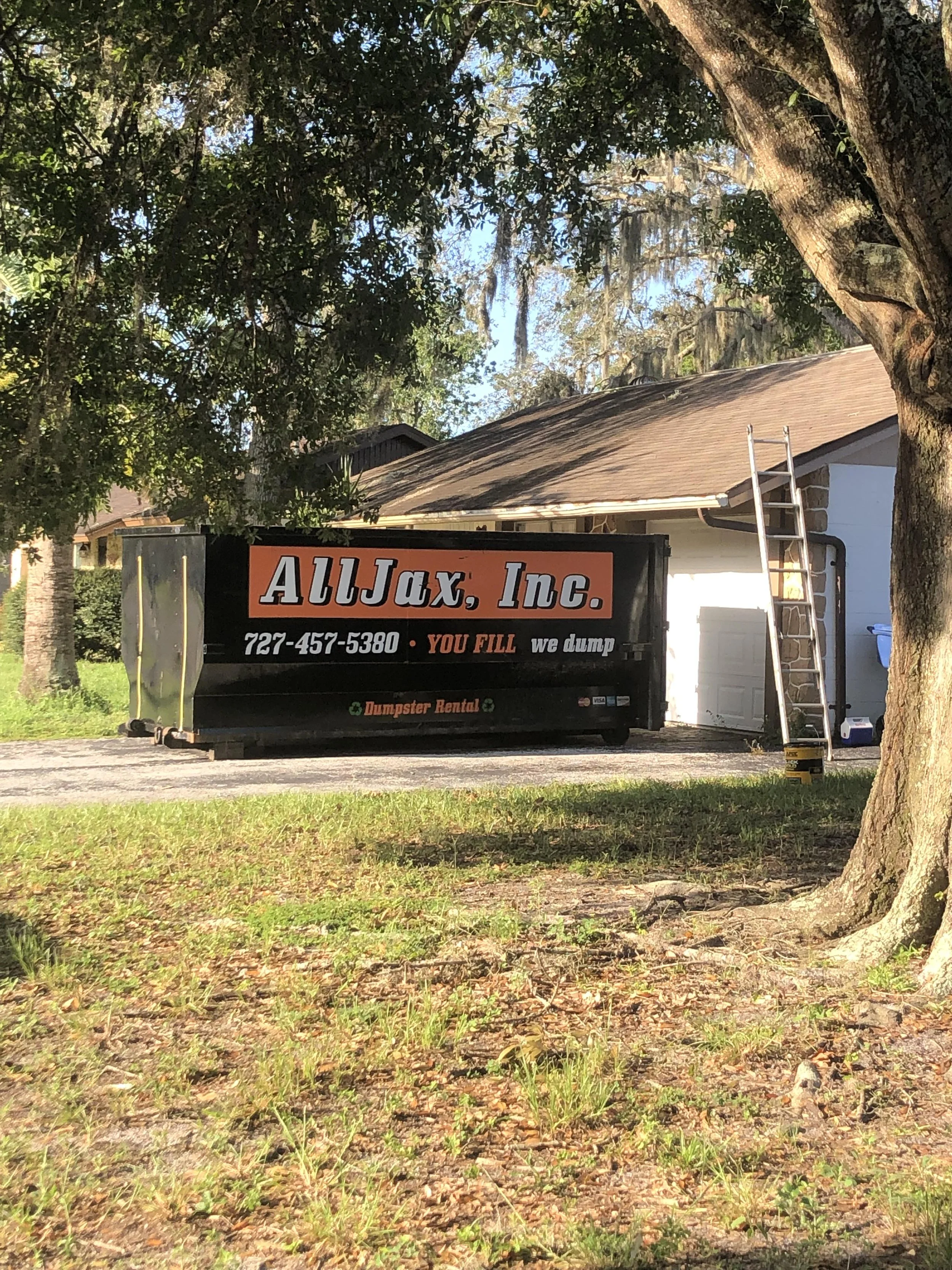 A house with a brown roof, a large tree in the front yard, and a Black dumpster with company branding and phone number in the driveway.