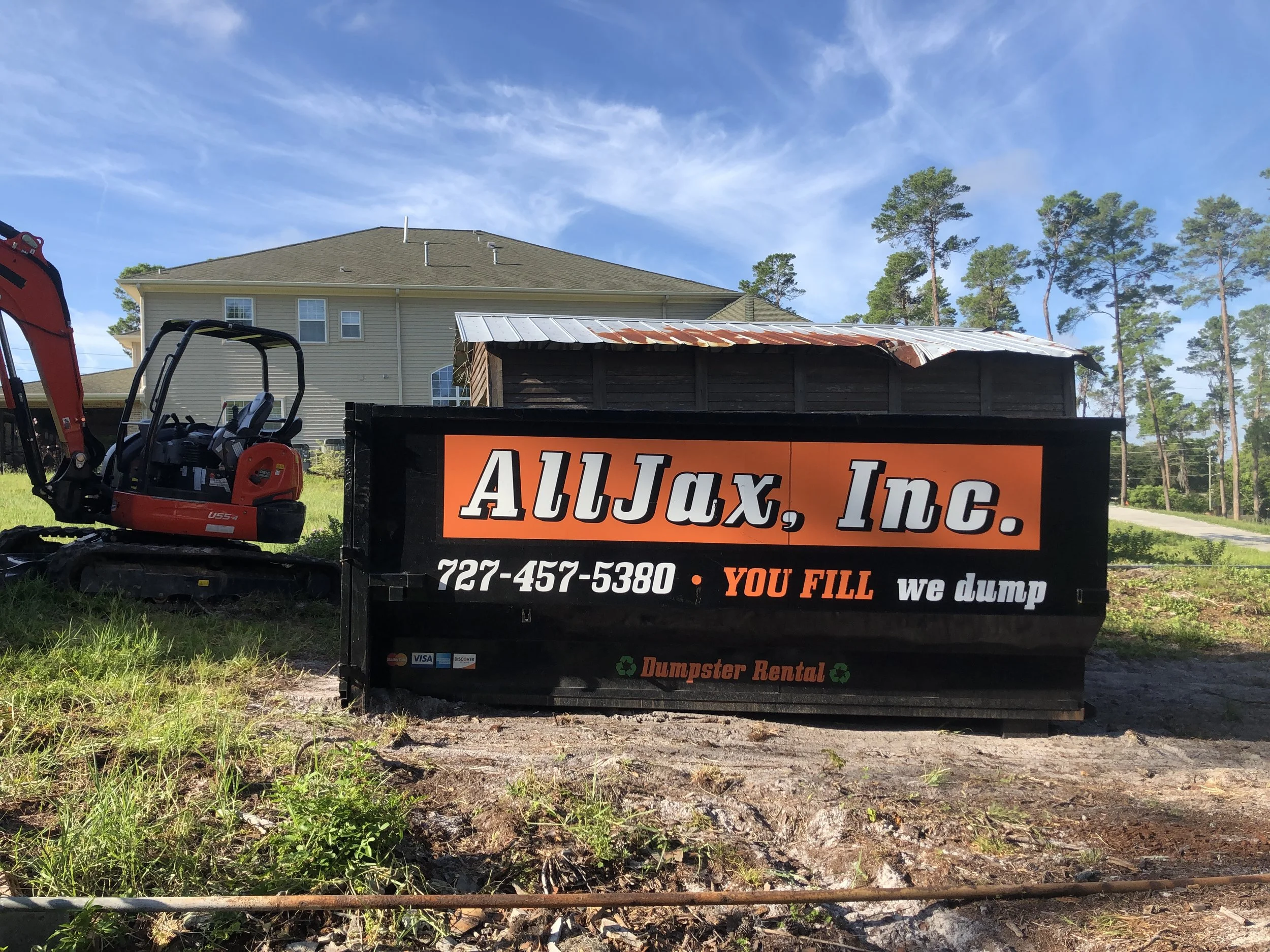 A small red and black excavator machine with a black dumpster with orange and white text, parked on green grass near a house with tan siding and multiple windows. The sky is partly cloudy with some tall trees in the background.