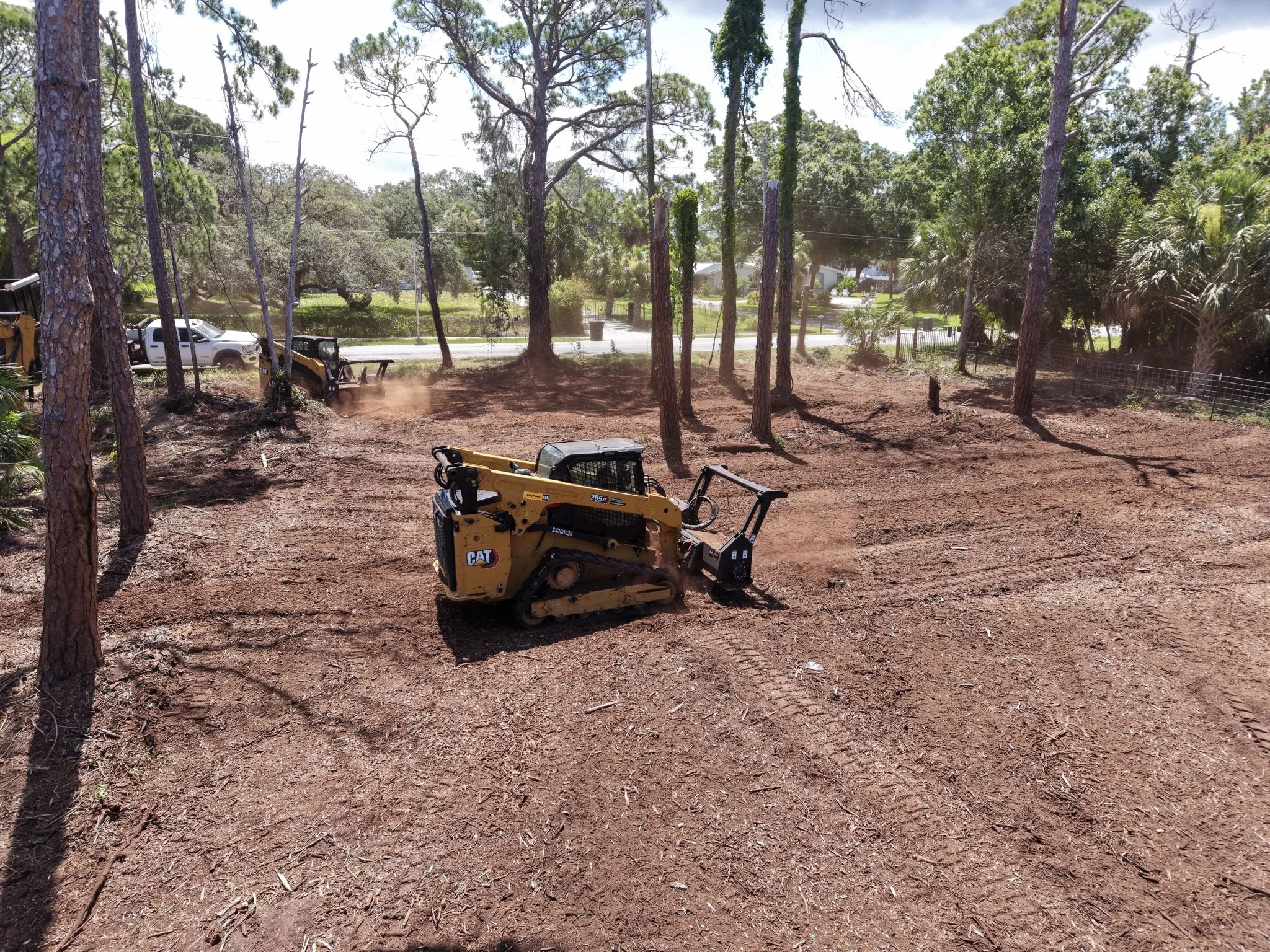 A small yellow excavator operating on a dirt clearing surrounded by trees, a white pickup truck parked nearby, and houses in the background.