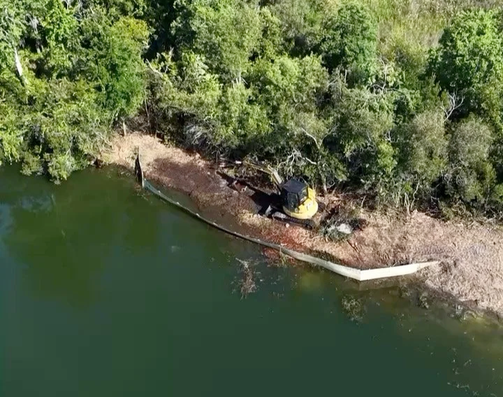A yellow construction vehicle and a pipe or barrier on a muddy shore beside a body of water, surrounded by green trees.