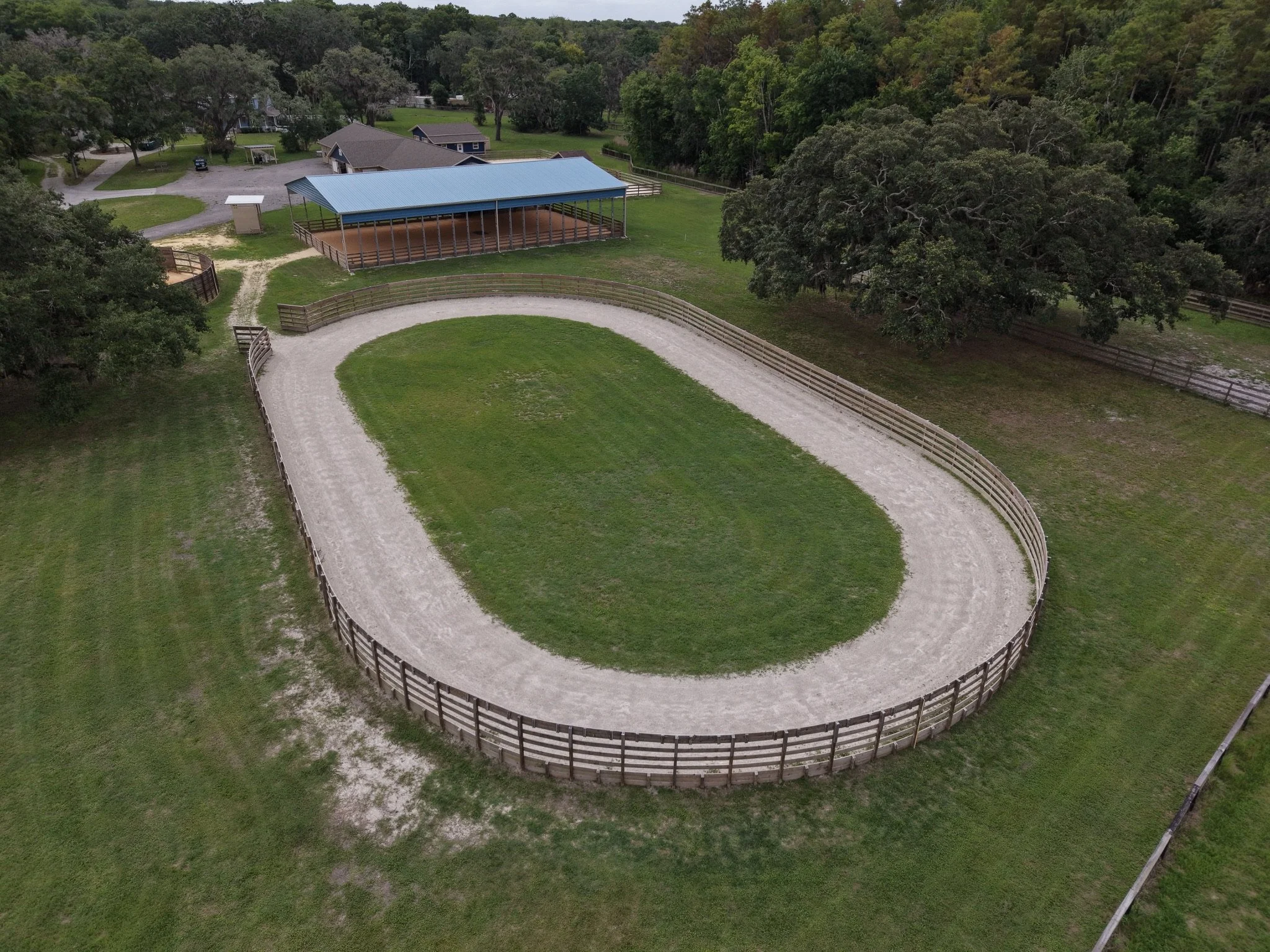 Aerial view of a horse riding ring with a curved wooden fence, surrounded by green grass and trees, with a covered shelter inside the ring and some small buildings nearby.