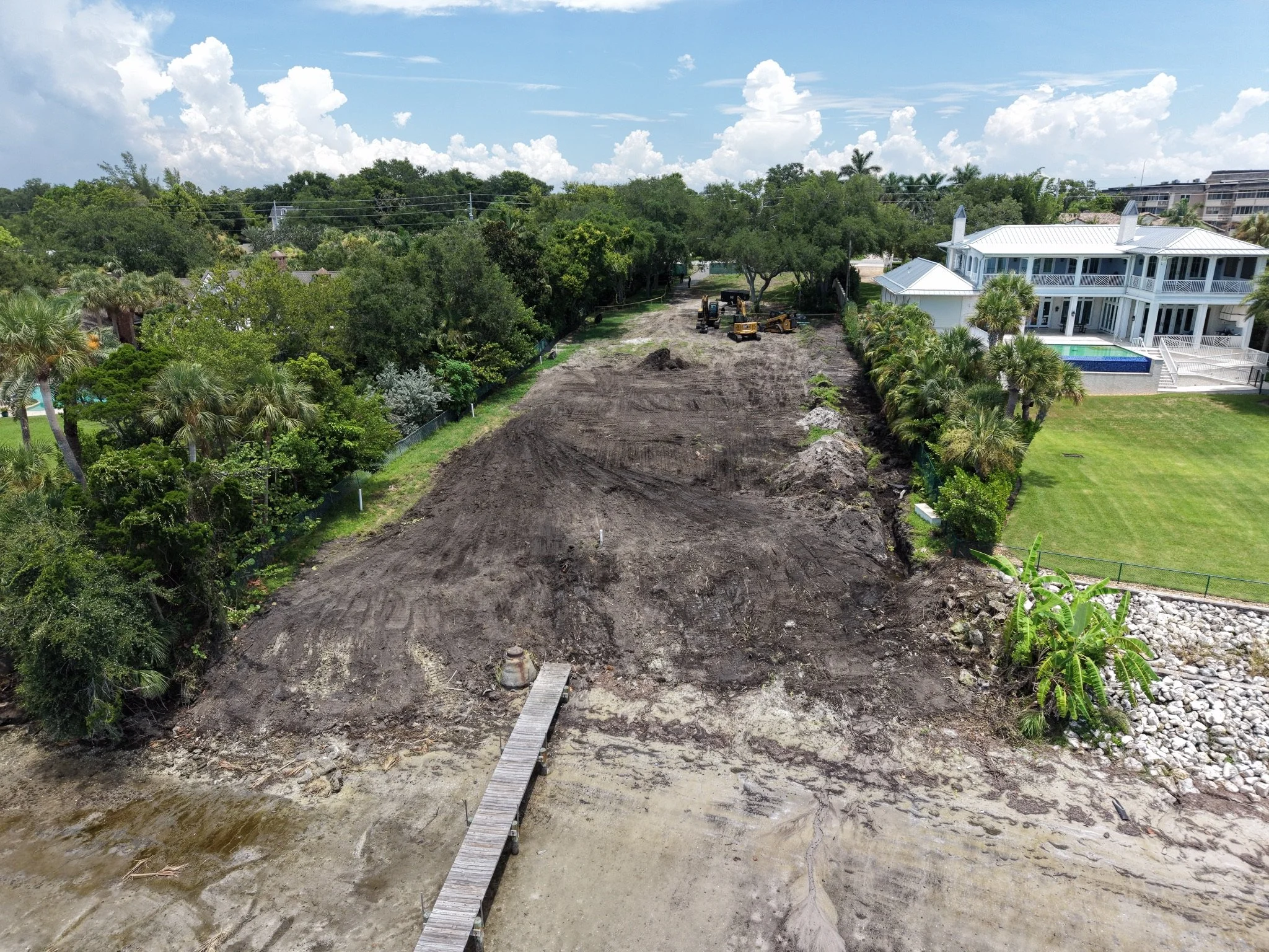 A cleared construction site with dirt and soil, a small wooden bridge over water, surrounded by trees and a neighboring white house with a porch and pool, under a partly cloudy sky.