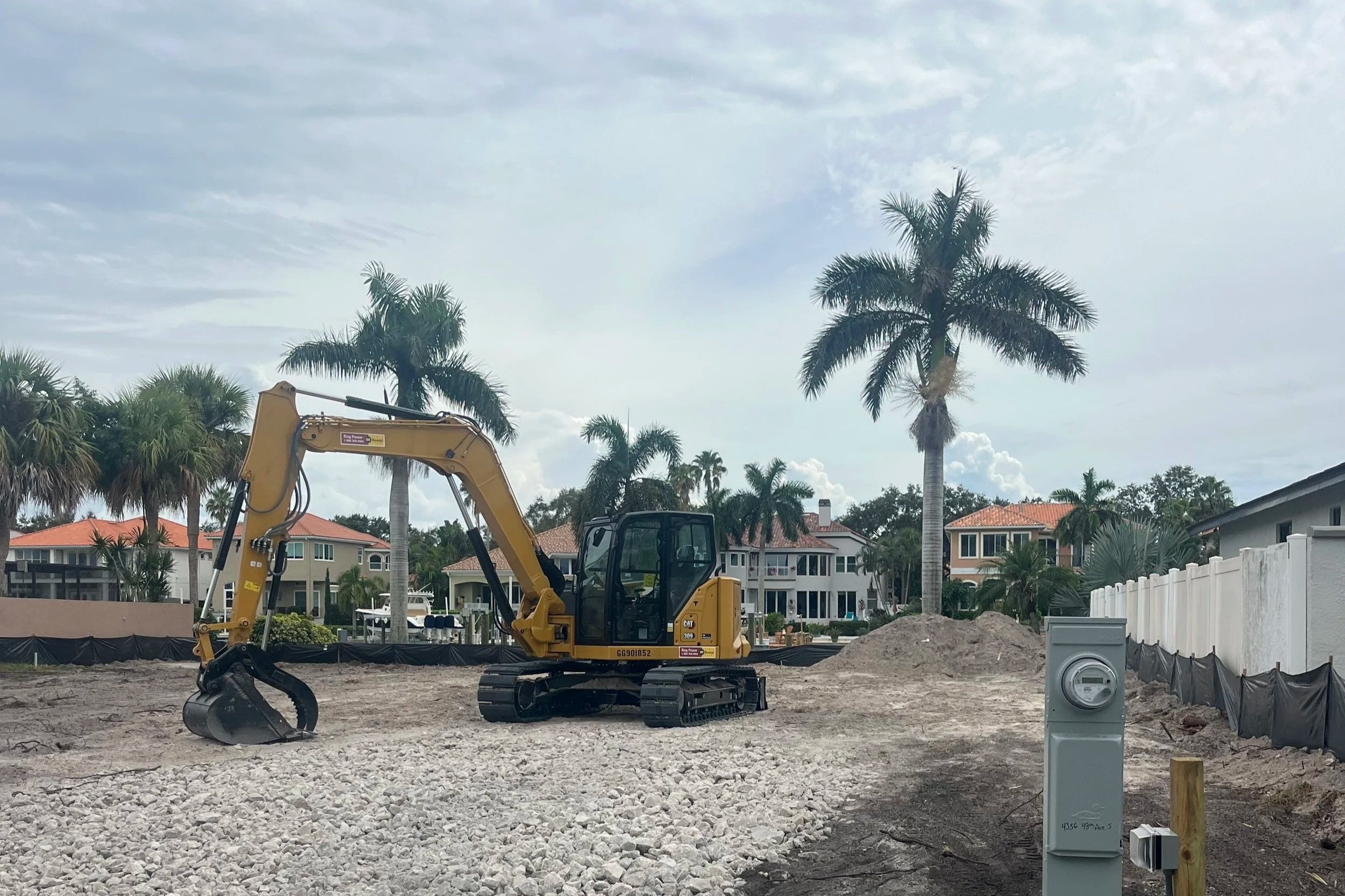 A construction site with a yellow excavator digging into the ground. In the background, there are tall palm trees and residential houses with red-tiled roofs. The sky is partly cloudy.