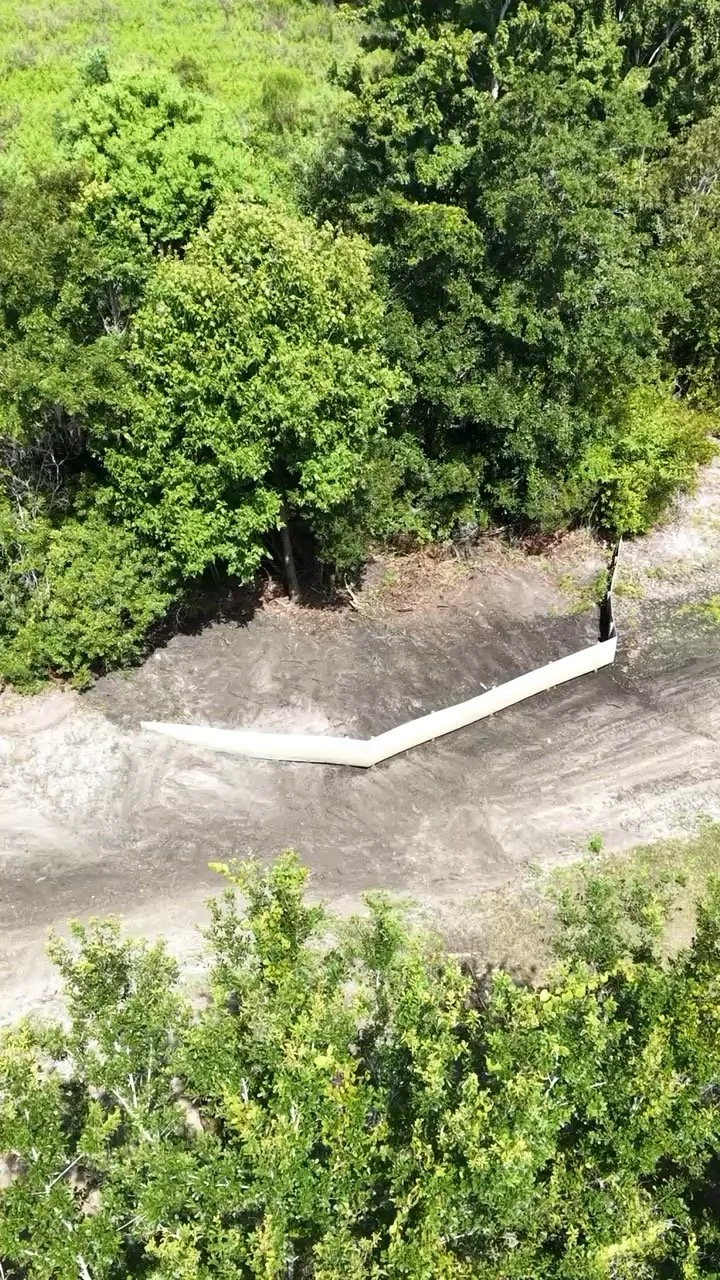 An aerial view of a dirt area with a white pipe or barrier running through it, surrounded by dense green trees.