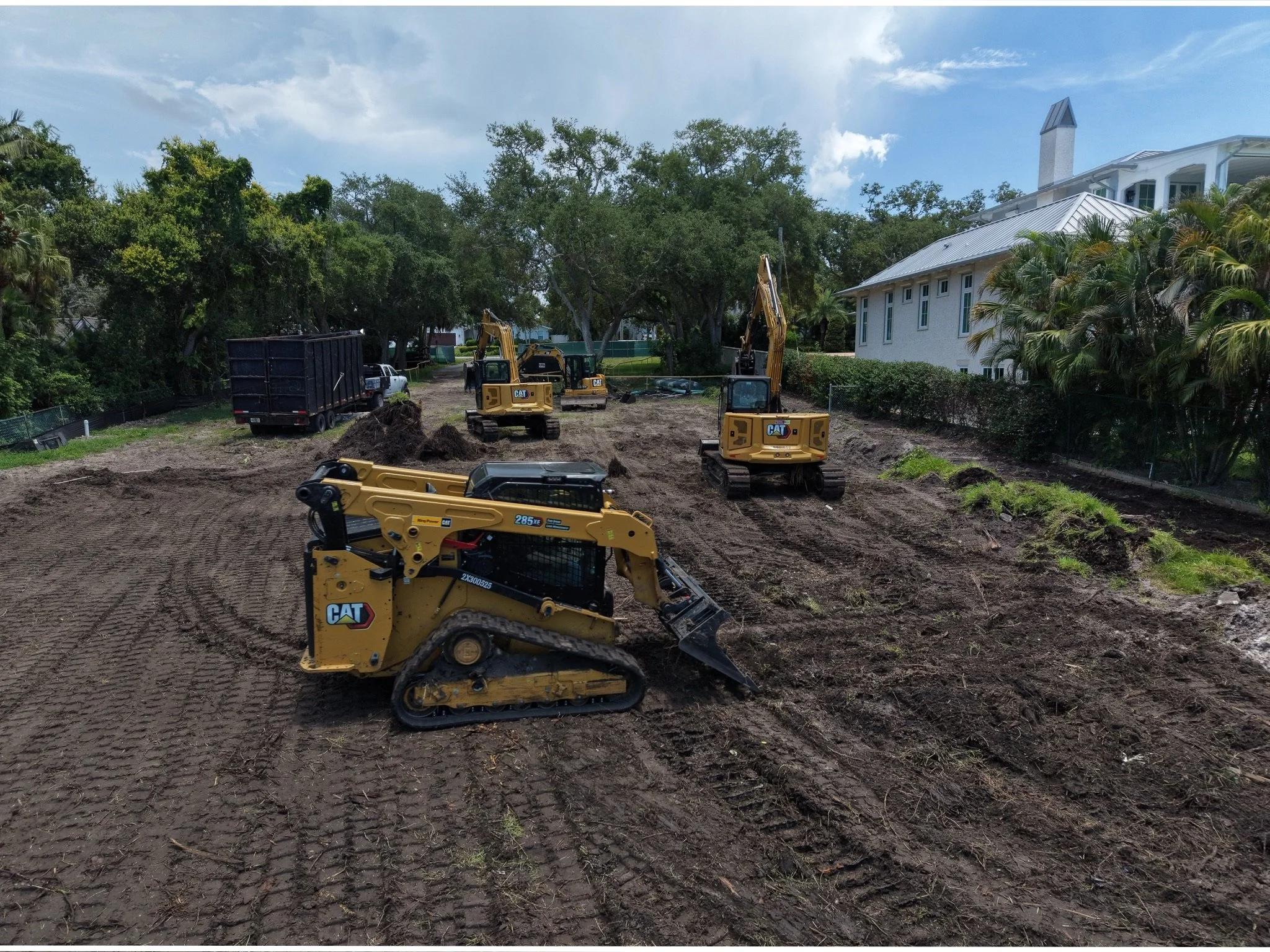 Construction site with excavators and a dump truck, with a building and trees in the background.