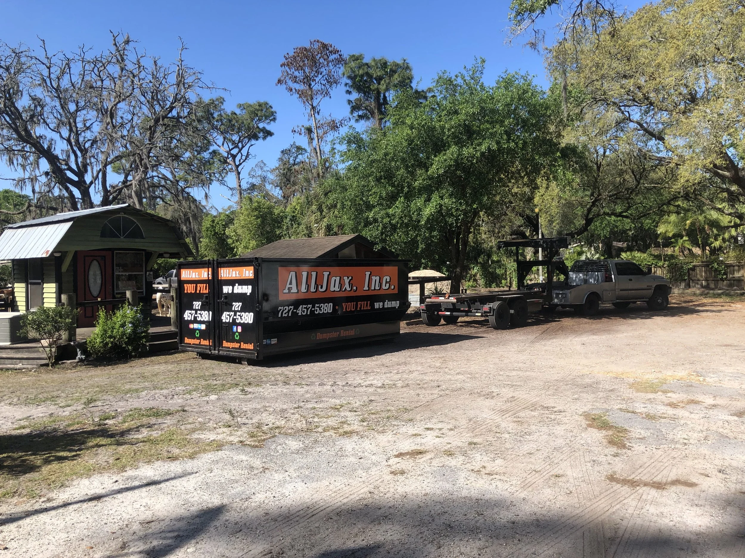 A property with trees and a small building at the front. There is a black dumpster labeled "AllJax, Inc" with a phone number and a truck parked nearby. The sky is clear and blue.