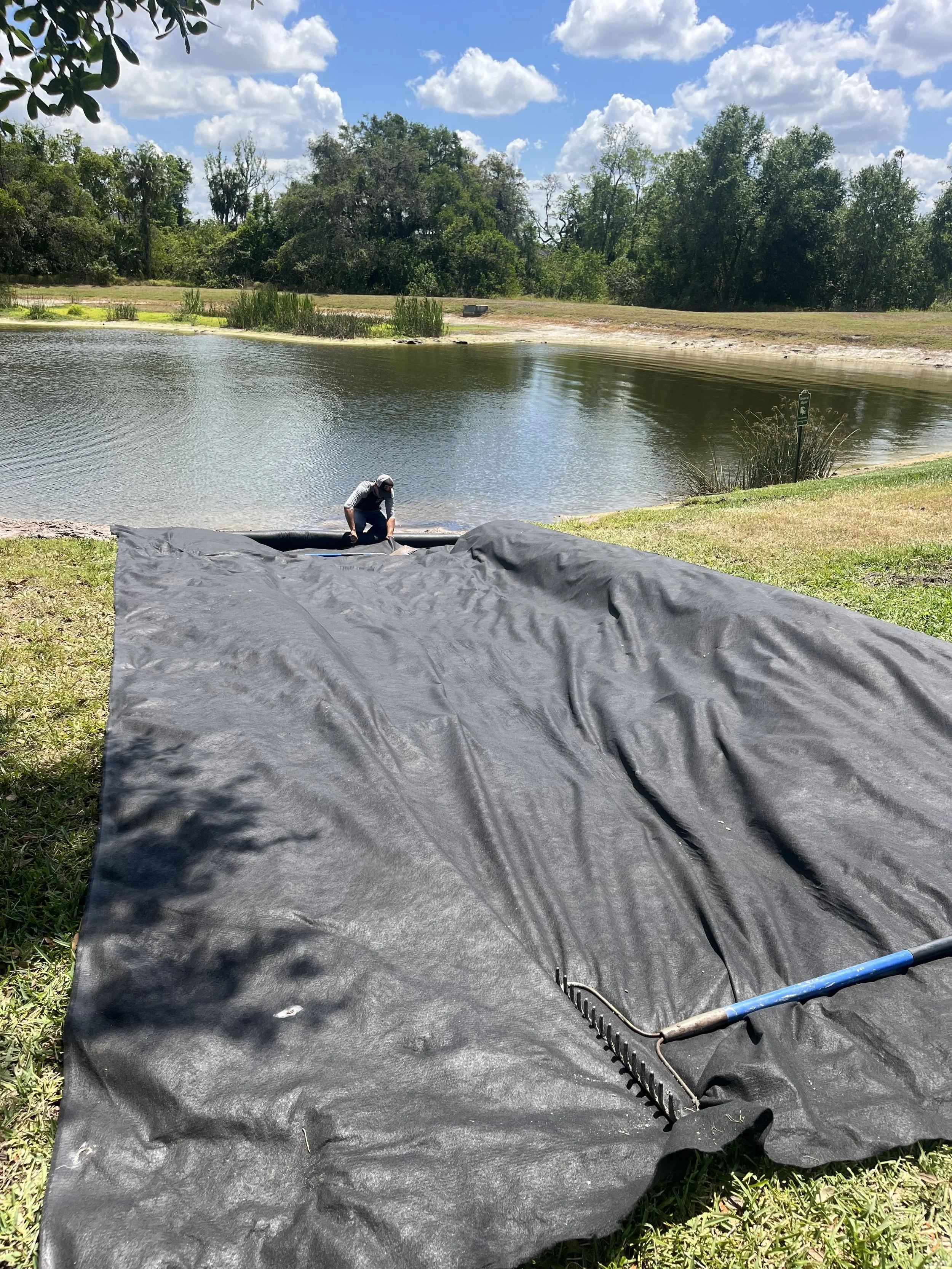 A person in a gray shirt and hat working on a black pond liner next to a lake, with grass, trees, and a partly cloudy sky in the background.