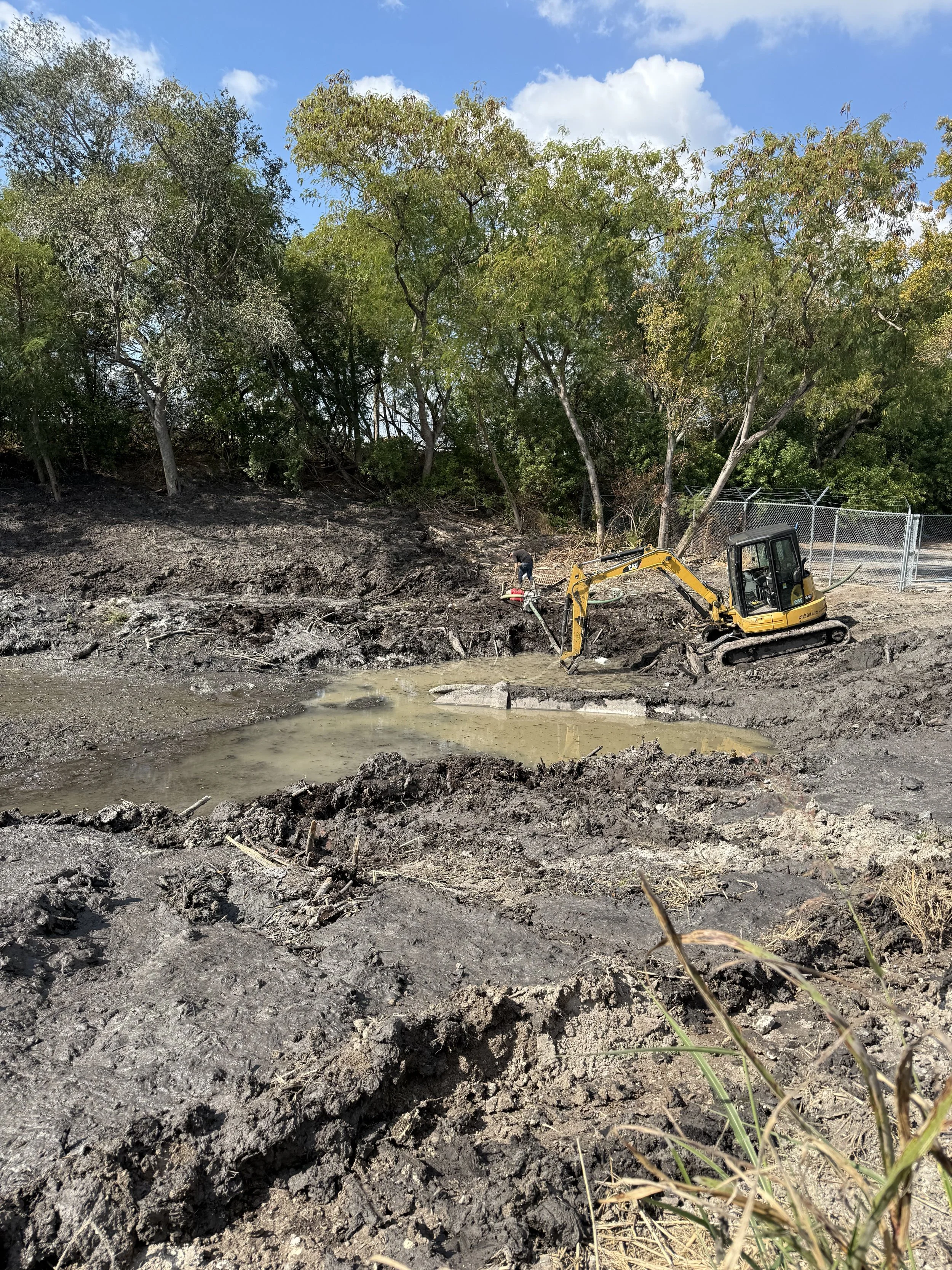 Construction site with a small yellow excavator on muddy ground near trees, work being done near a fenced area.