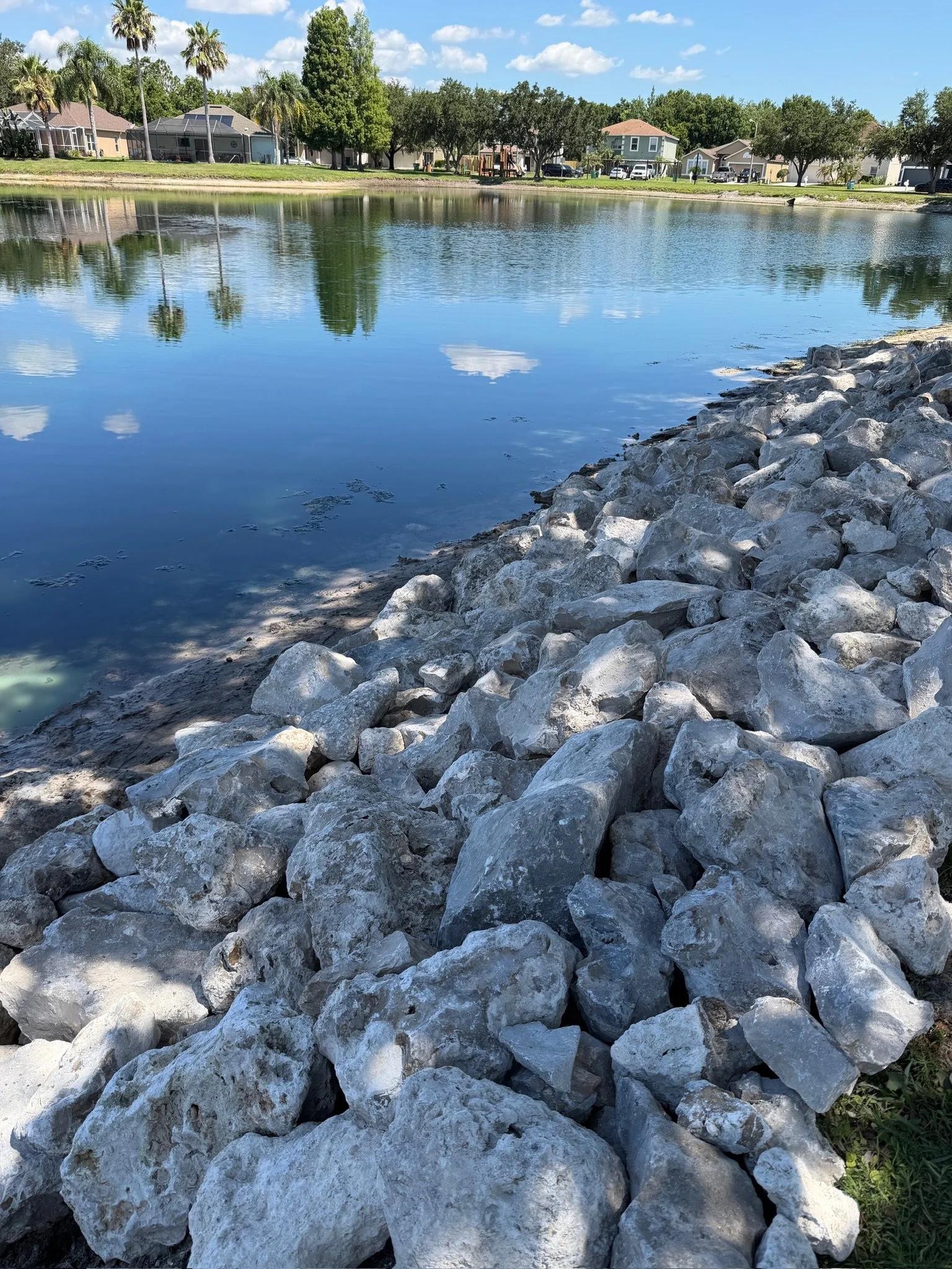 View of a lakeside scene with large rocks in the foreground, calm water reflecting trees and houses across the lake, some small clouds in the blue sky.