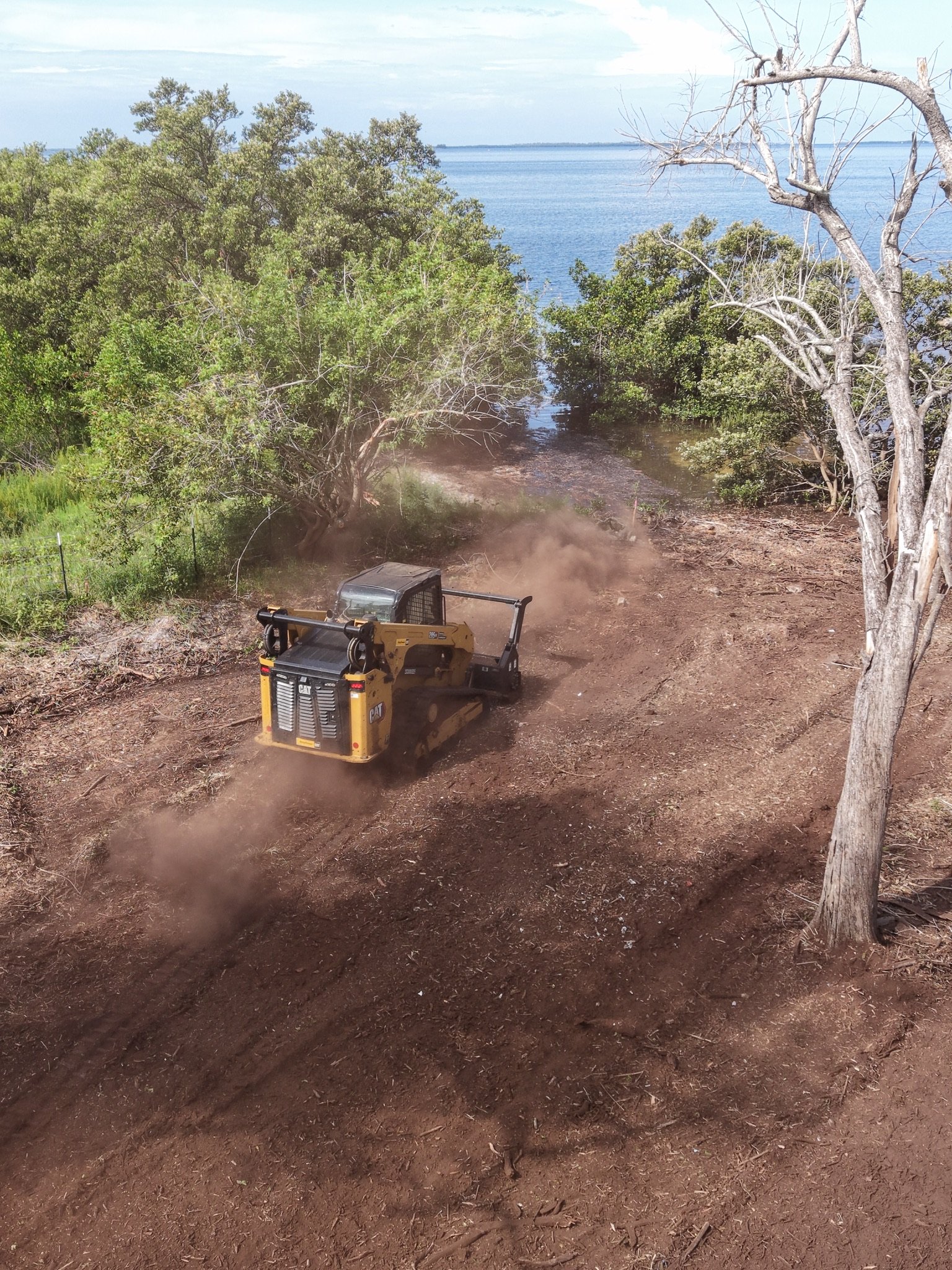A construction vehicle operating on a dirt path near a body of water, with trees and shrubs along the shoreline and a cloudy sky above.