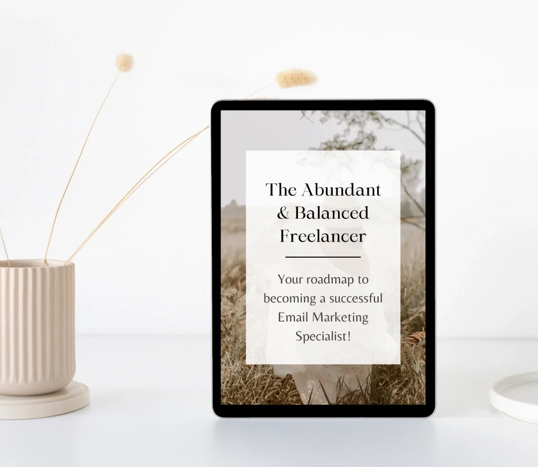 An electronic tablet displays a cover for a guide titled 'The Abundant & Balanced Freelancer,' aimed at becoming a successful email marketing specialist. The tablet is on a white surface next to a beige, ribbed ceramic vase with dried flowers and a white round dish.
