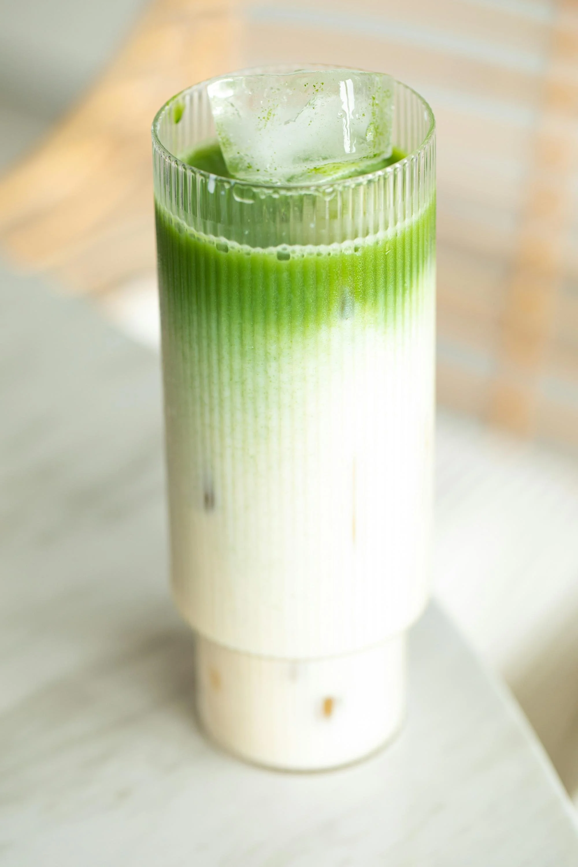 A glass of green matcha latte with ice cubes on top, placed on a light-colored surface.