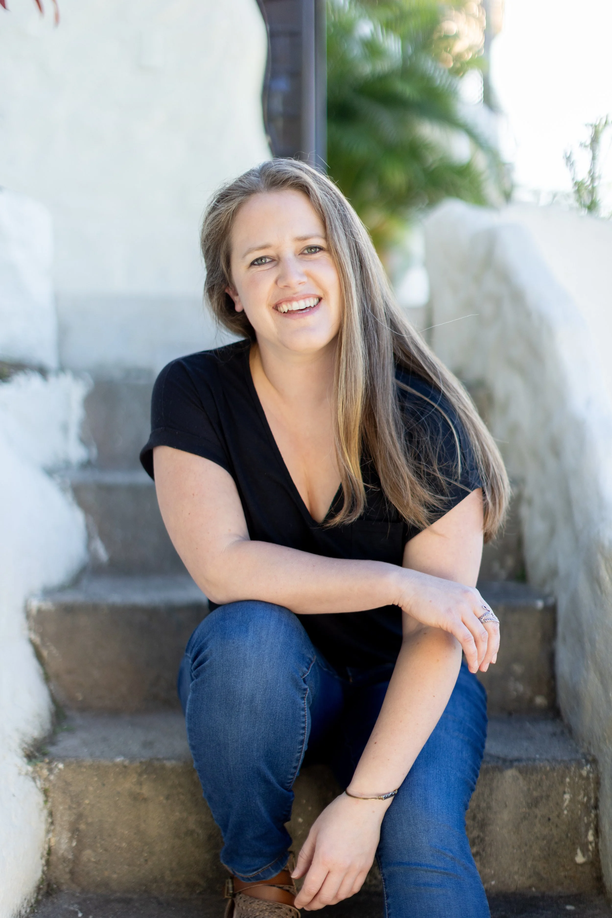 A woman with long brown hair seated on outdoor steps, wearing a black top and blue jeans, smiling at the camera with arms crossed and surrounded by white stone walls and greenery.