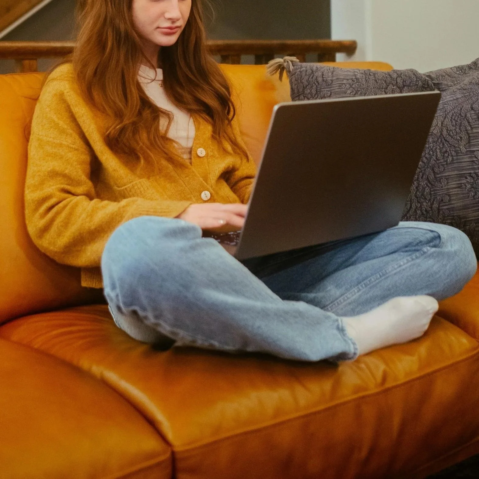 A person with long red hair wearing a yellow sweater, white shirt, and jeans is sitting on a brown leather couch using a laptop. They are barefoot and sitting cross-legged. There are decorative cushions behind them.