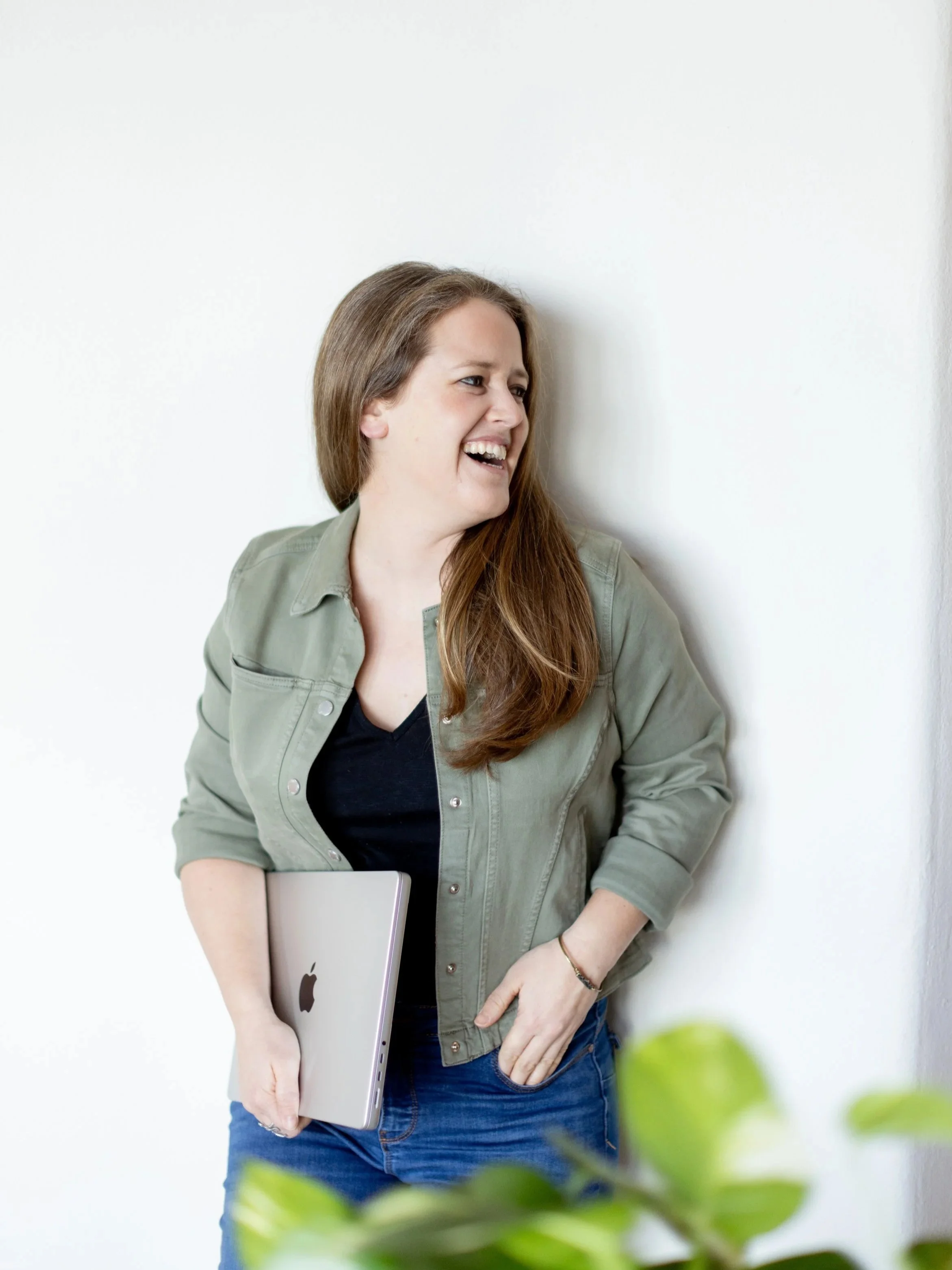 A woman with long brown hair smiling and leaning against a white wall, holding a closed laptop in her left hand, with blurred green leaves in the foreground.