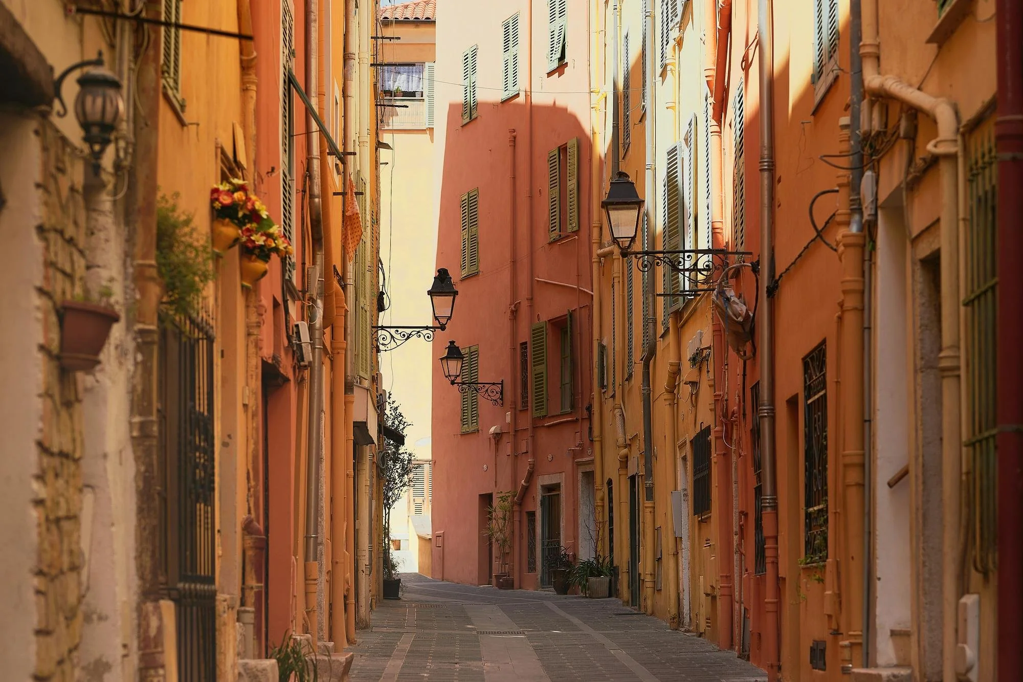 A narrow alleyway with colorful Mediterranean-style buildings on both sides. The buildings are painted in shades of orange, yellow, and peach, with green shutters on the windows. Several street lamps hang across the alley, and small potted plants are visible near the windows. The alley is paved with stones and appears to be in a quaint, sunny European neighborhood.