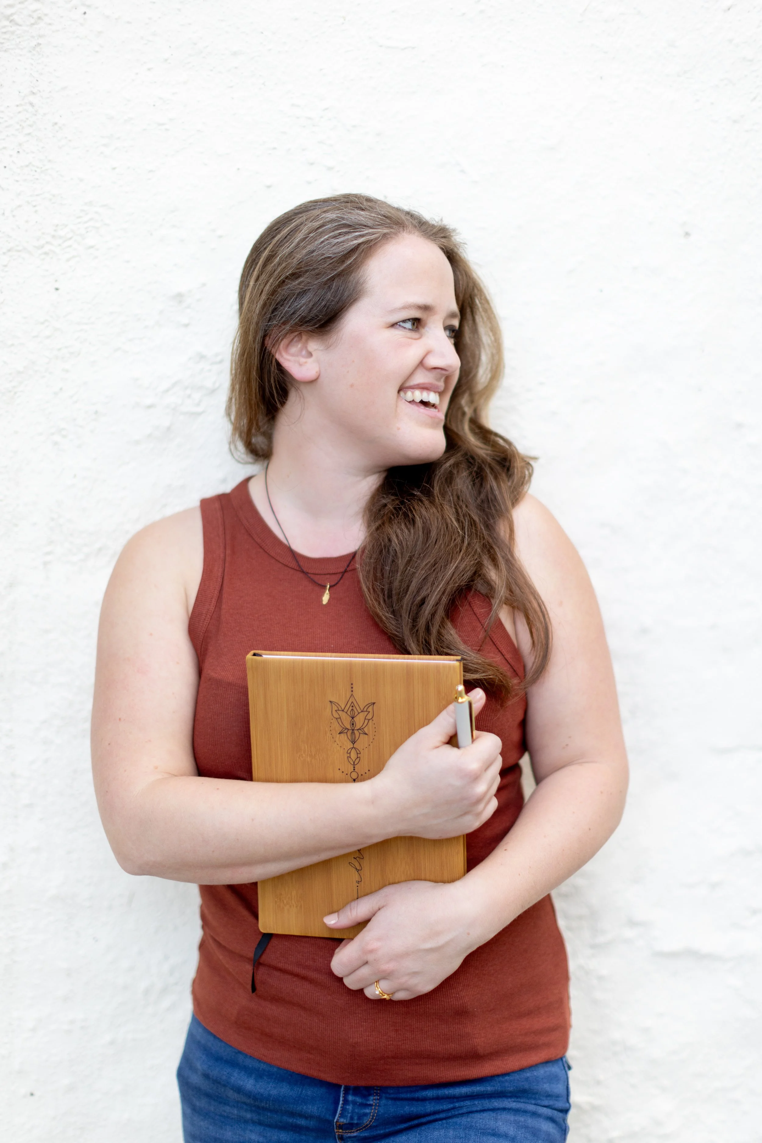 A woman with long brown hair and light skin, wearing a sleeveless reddish-brown top, blue jeans, and a delicate gold necklace, smiling while holding a wooden notebook or journal with an engraved floral design and a pen in front of a white textured wall.