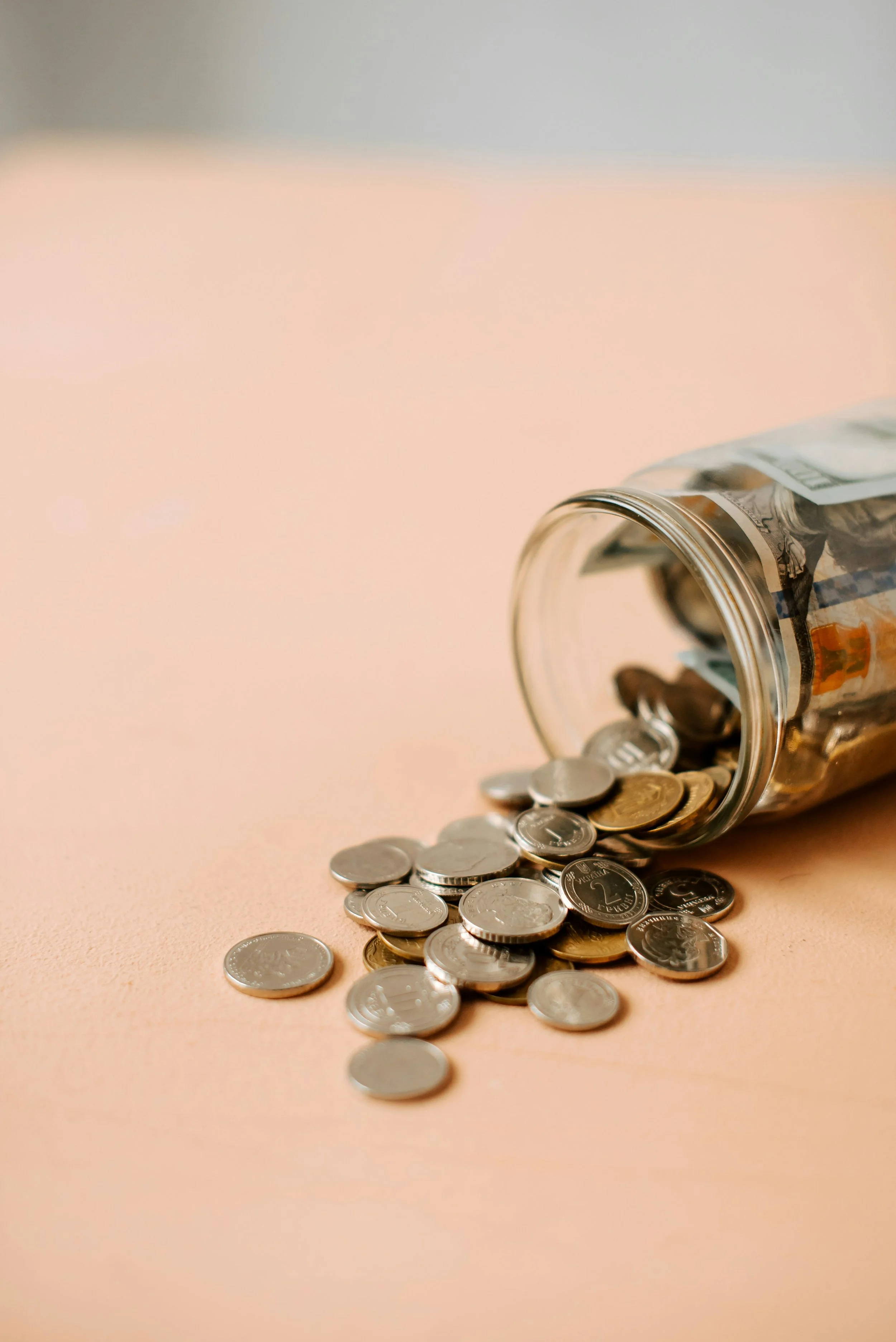 A glass jar tipped over with coins spilling onto a beige surface.