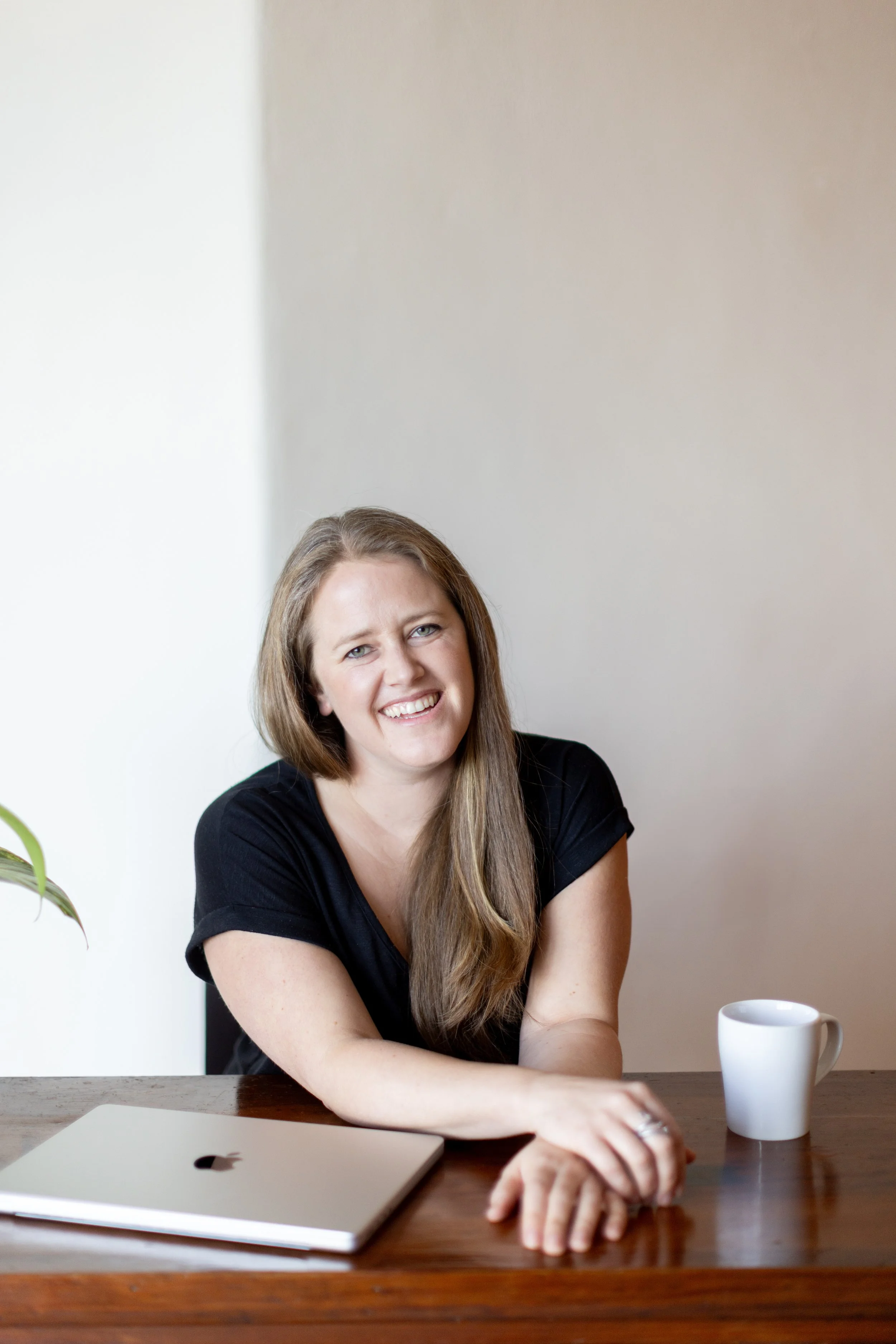A woman with long brown hair smiling, sitting at a wooden table with a silver laptop and a white mug, in a minimalistic room with white walls and a plant partially visible on the left.