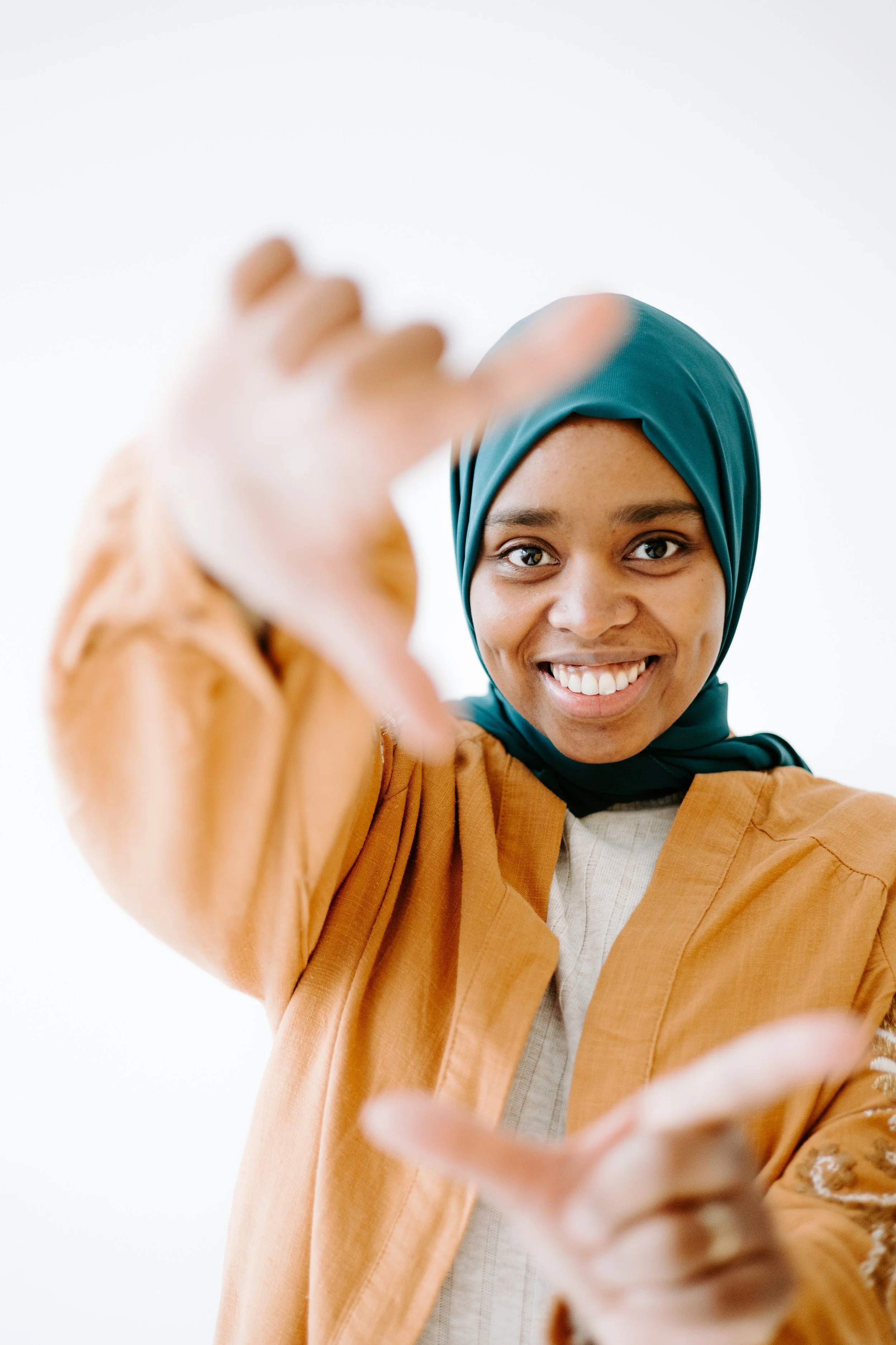 A smiling woman wearing a teal headscarf and brown jacket, reaching towards the camera with her hands.