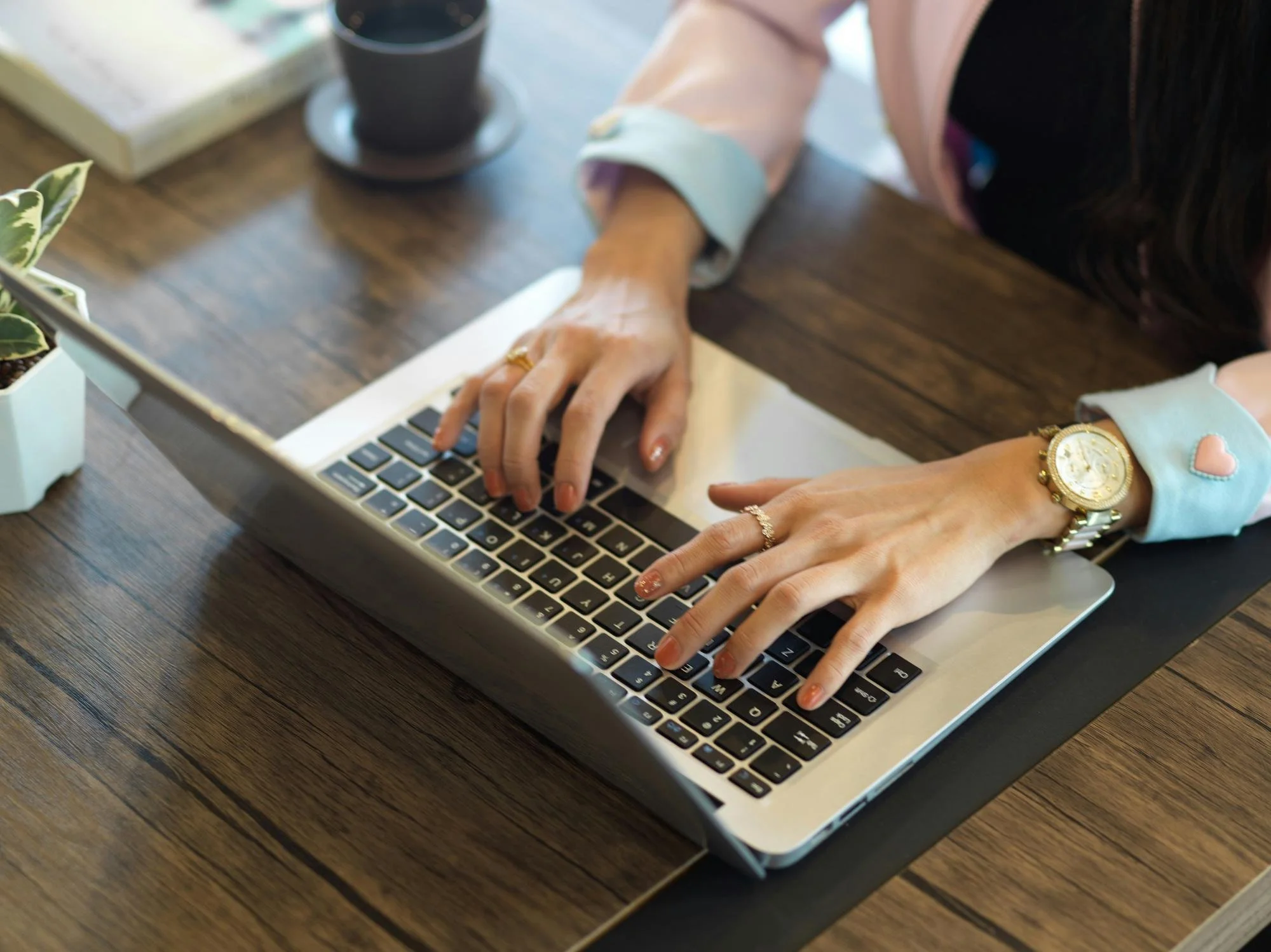 A person is wearing a pink blazer and a gold watch, the other has colorful nails and a gold ring typing on a MacBook laptop computer at a wooden table. There is a small potted plant and a cup of coffee or tea on the table.