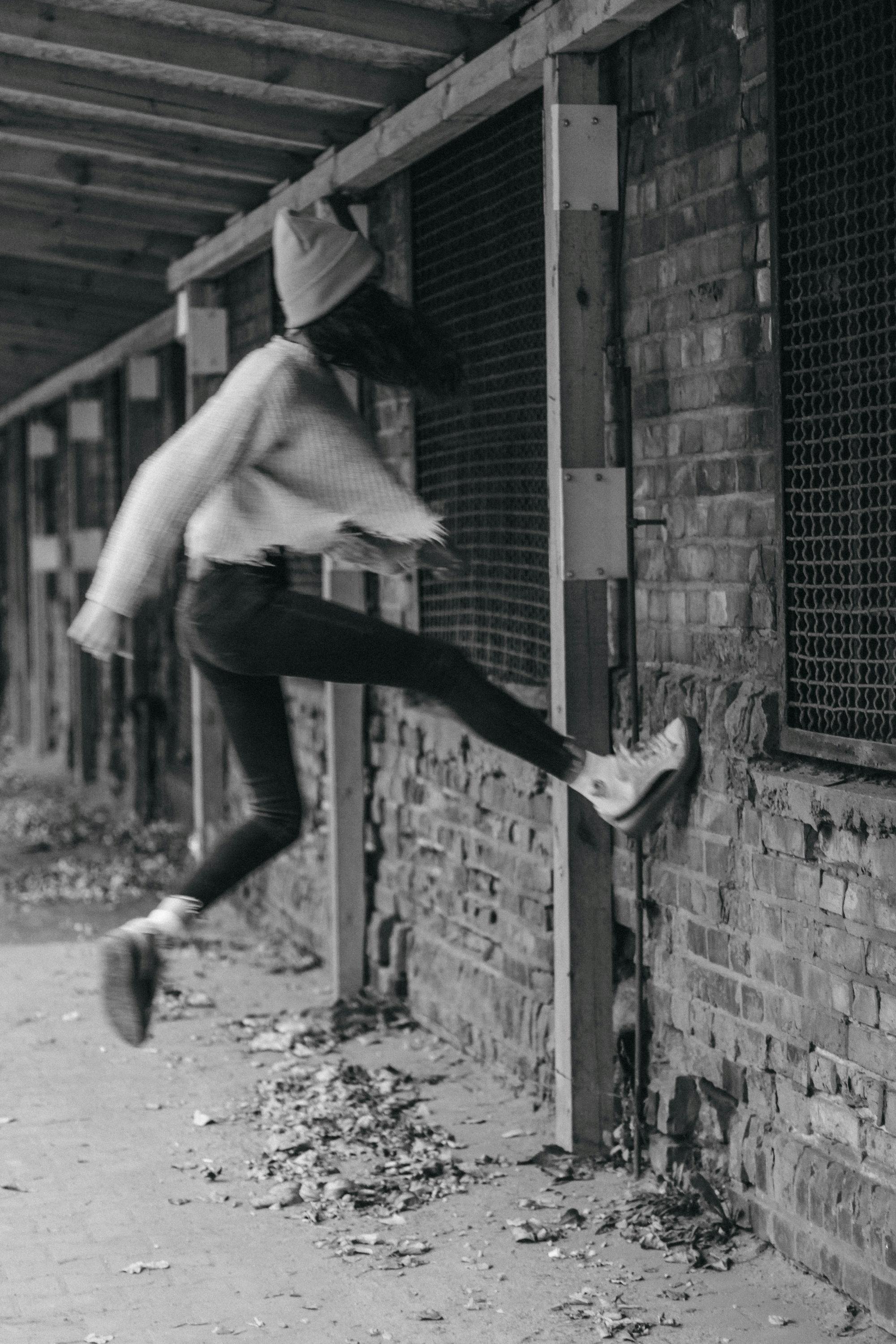 A person in a light-colored jacket, beanie, and sneakers is jumping against a brick wall with a grated window, in an outdoor area with fallen leaves on the ground.