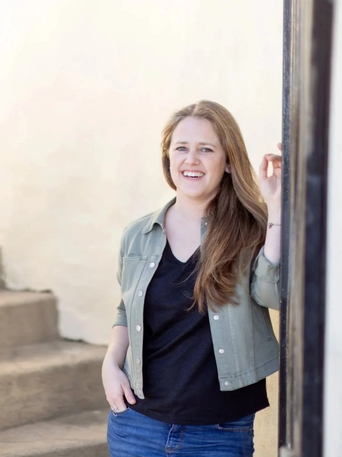 A woman with long red hair, wearing a green jacket over a black shirt, smiling and standing against a beige wall, partially leaning on a black vertical surface.