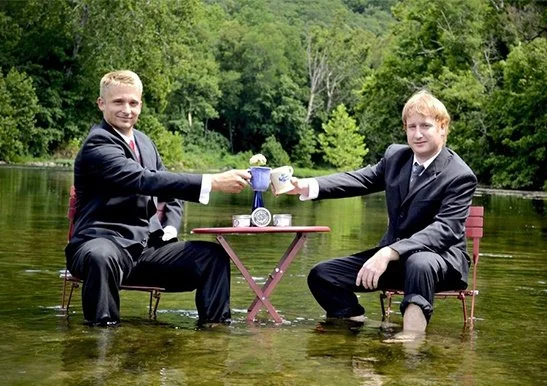 Two men in suits sitting in a river, having a tea party with a small table and cups, surrounded by green trees.
