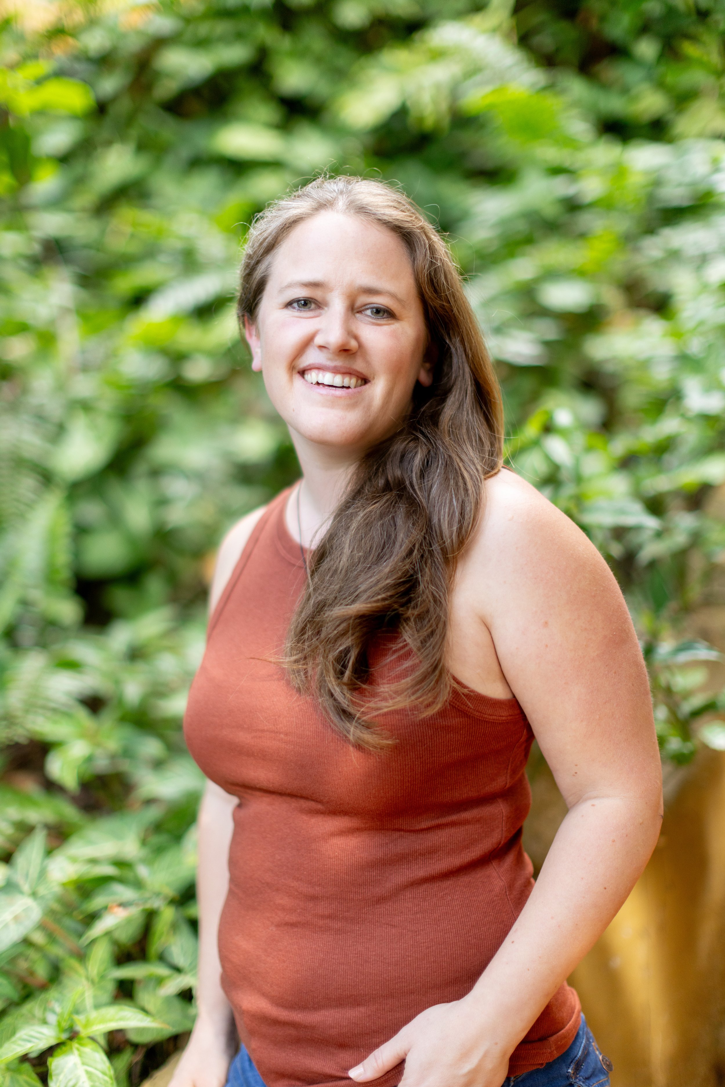 A woman with long brown hair, wearing a rust-colored tank top and blue jeans, smiling in front of a green leafy background.