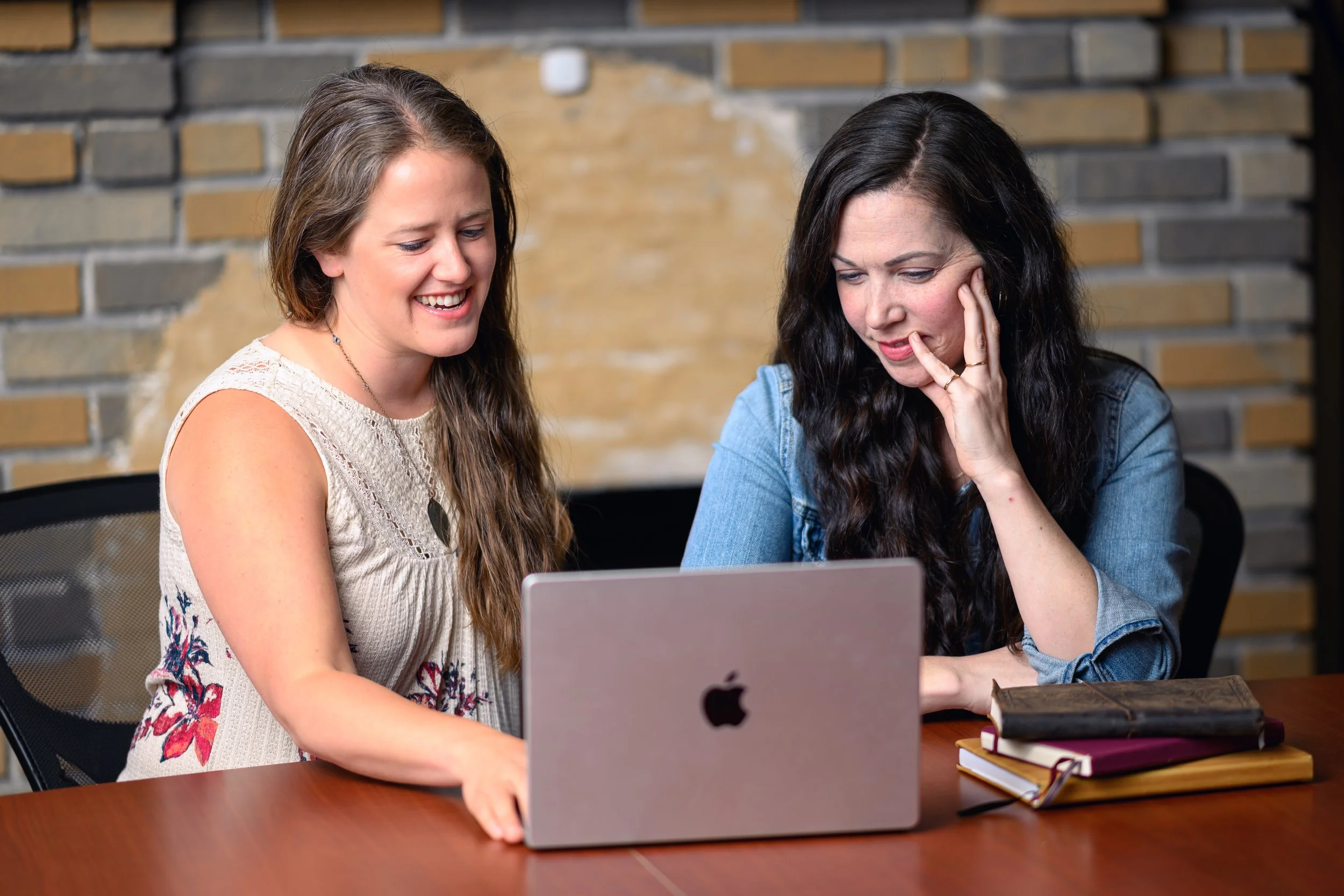 Two women sitting at a wooden table, looking at a laptop and discussing, with a brick wall in the background.