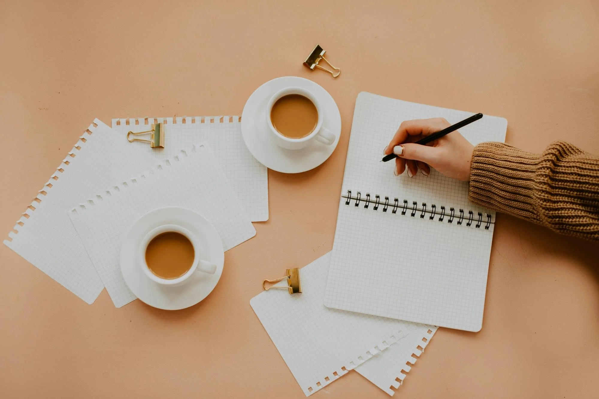 A flat lay of two cups of coffee, a person's hand writing in a graph paper notebook, three sheets of graph paper, four gold binder clips, and two gold clips on a peach-colored background.