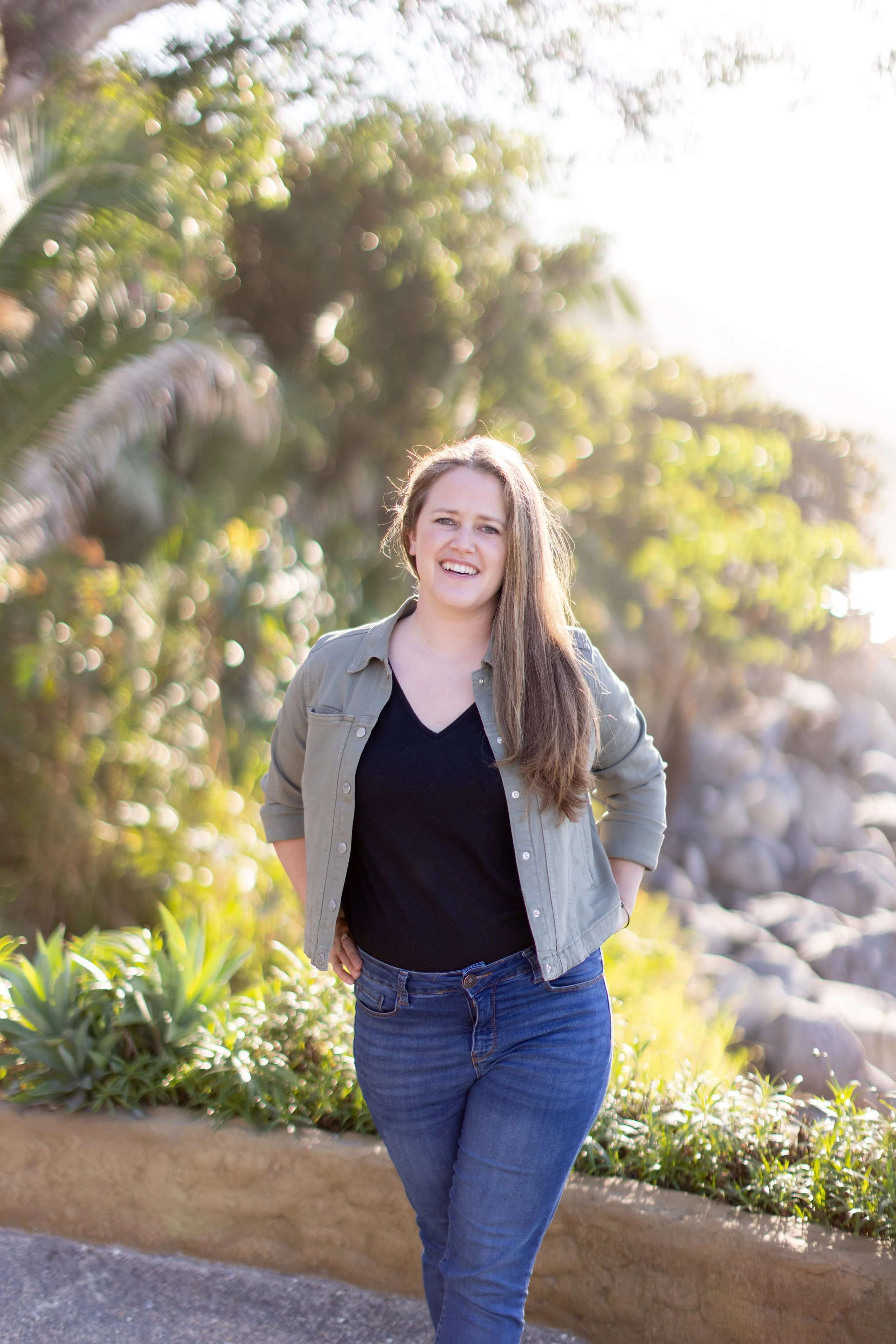 A young woman with long brown hair, wearing a black top, light green jacket, and blue jeans, standing outdoors on a sunny day with greenery and rocks in the background.