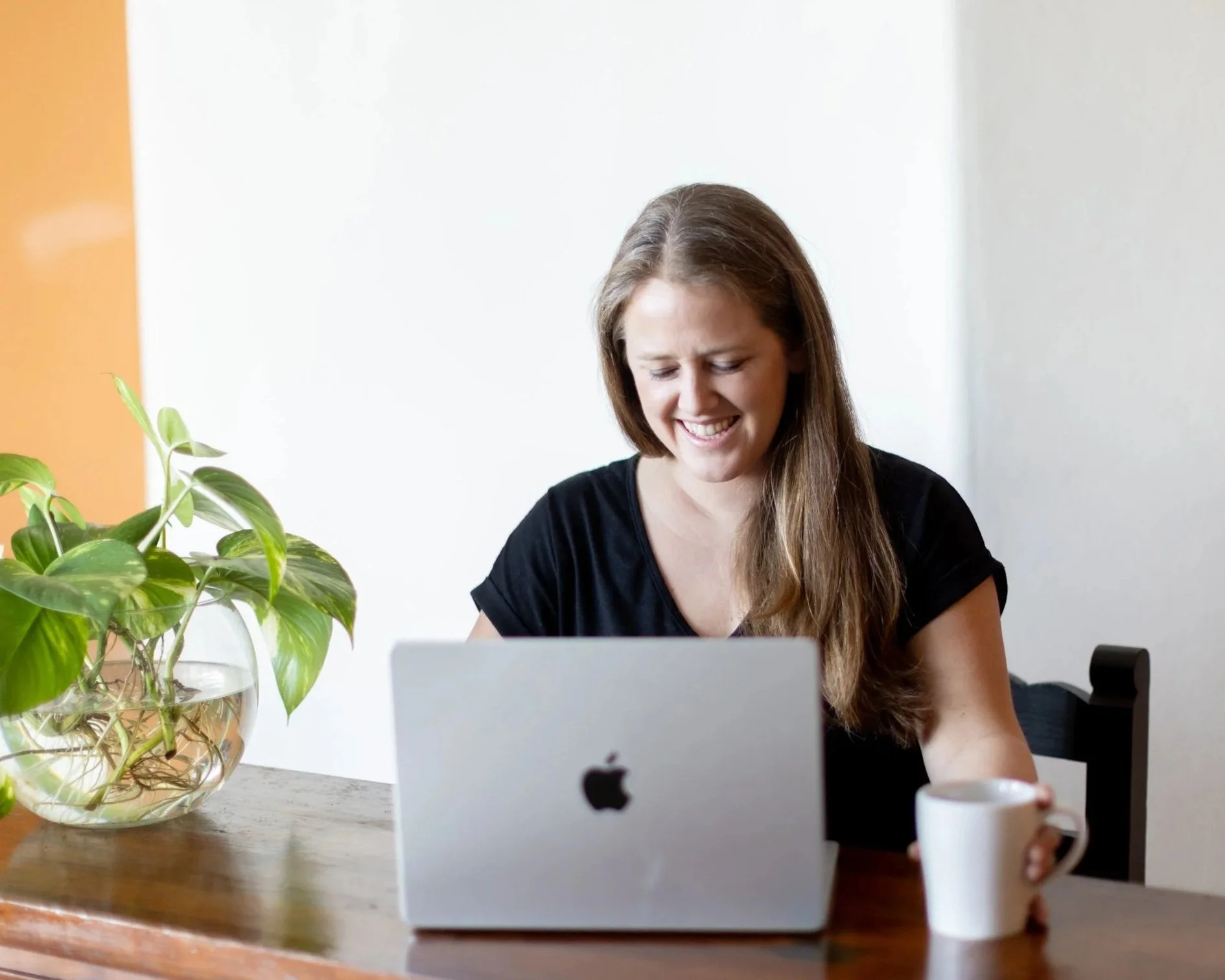 A woman with long brown hair smiling at her laptop on a wooden table, with a potted plant and a white coffee mug nearby.