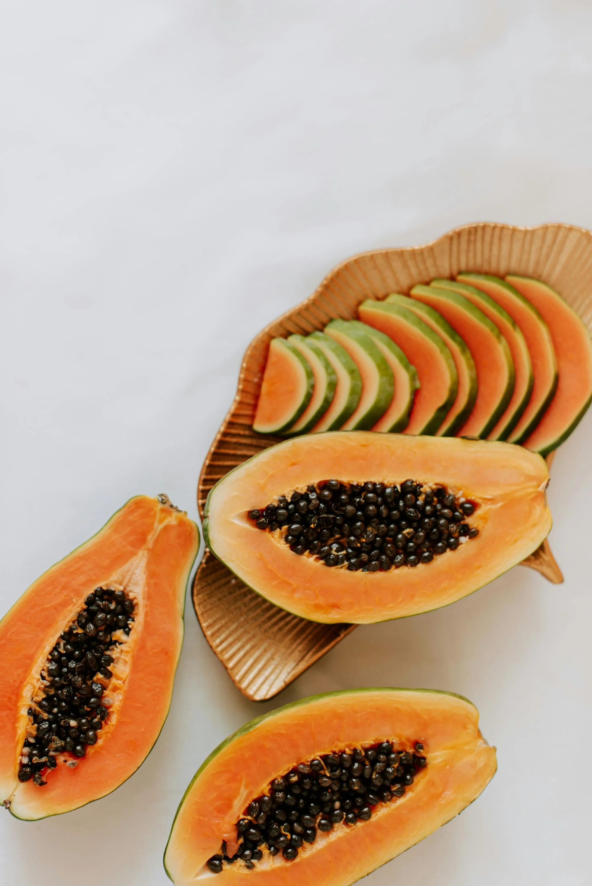 Slices of papaya with orange flesh and black seeds arranged on a wooden plate and scattered on a white surface.