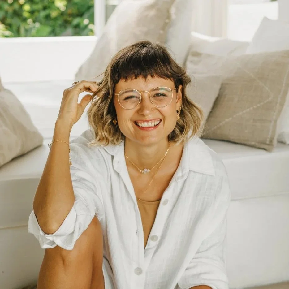 A woman with short, wavy hair and glasses smiling while sitting on a white sofa with beige pillows, in a bright room with windows and greenery outside.