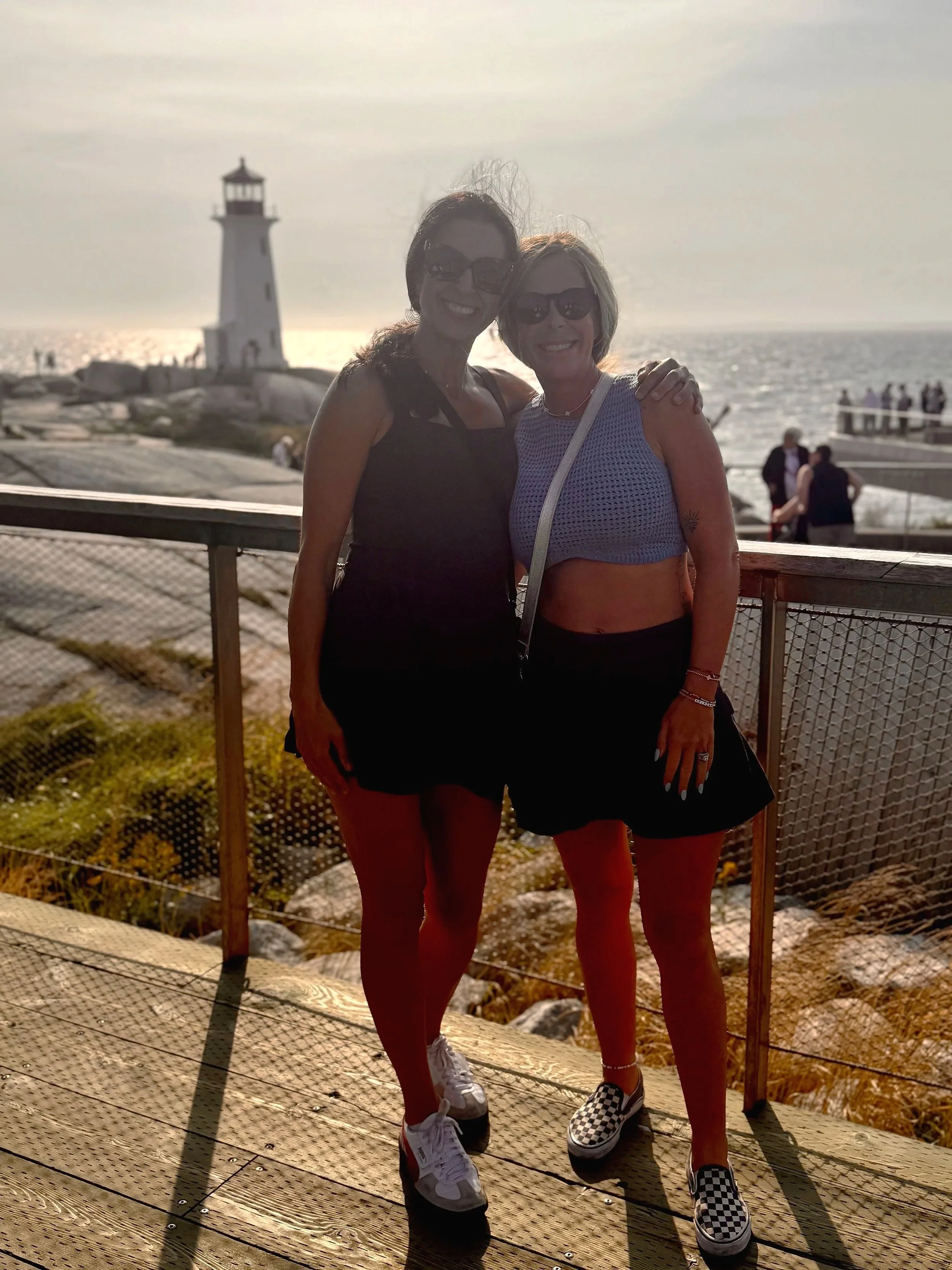 Two women standing together outdoors near a beach with a lighthouse in the background, smiling, wearing sunglasses, and casual summer clothes.