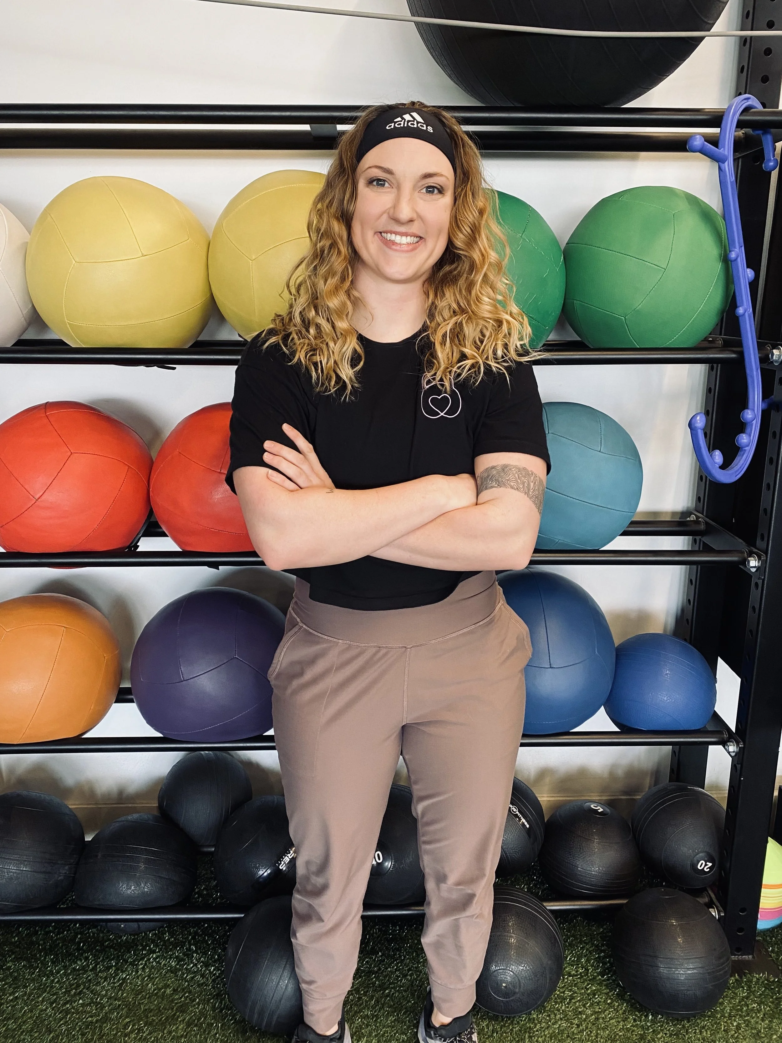 A woman with curly blonde hair, wearing a black Adidas headband and black t-shirt, stands with arms crossed in front of a rack of colorful exercise balls at a gym.