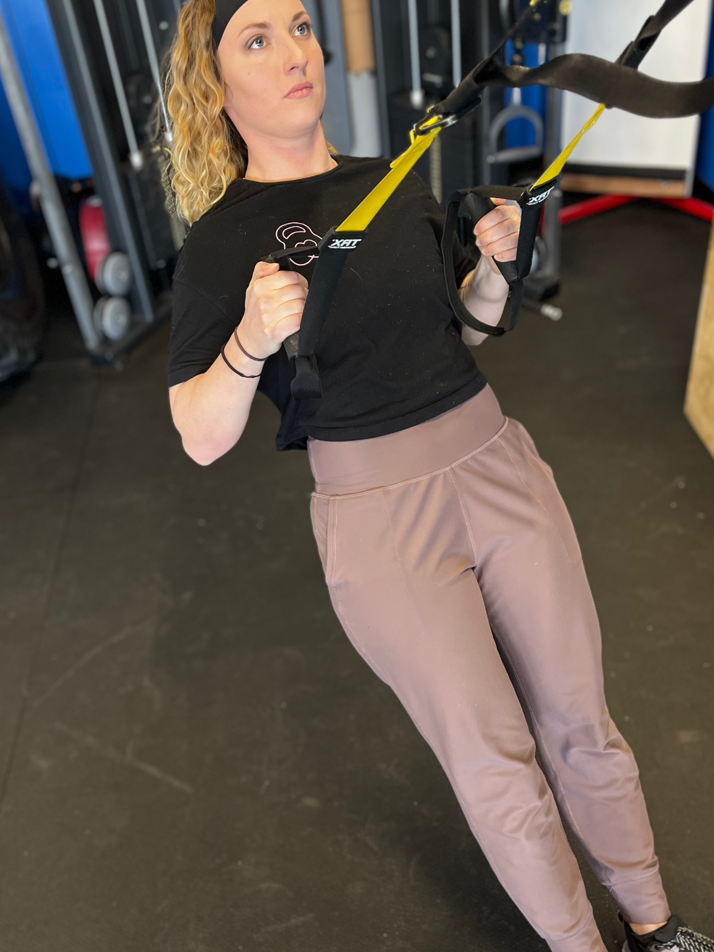 A woman with long blonde curly hair wearing a black shirt and beige pants using suspension training straps for exercise in a gym.