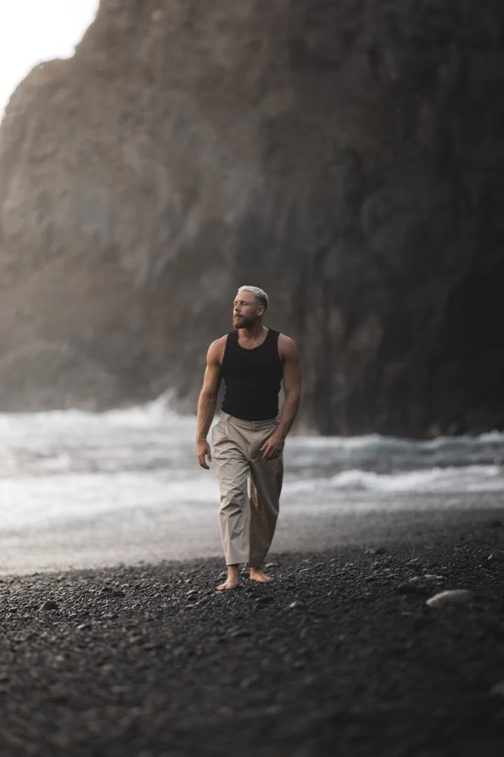 Tom Wilson-Leonard walking barefoot on a dark volcanic beach beneath coastal cliffs.