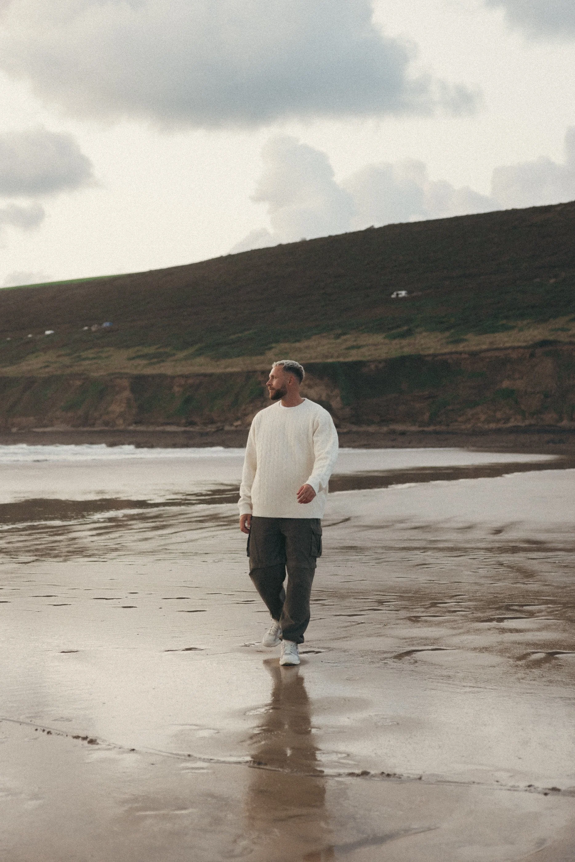 Tom walking along the shoreline in a cream jumper beneath an overcast sky.